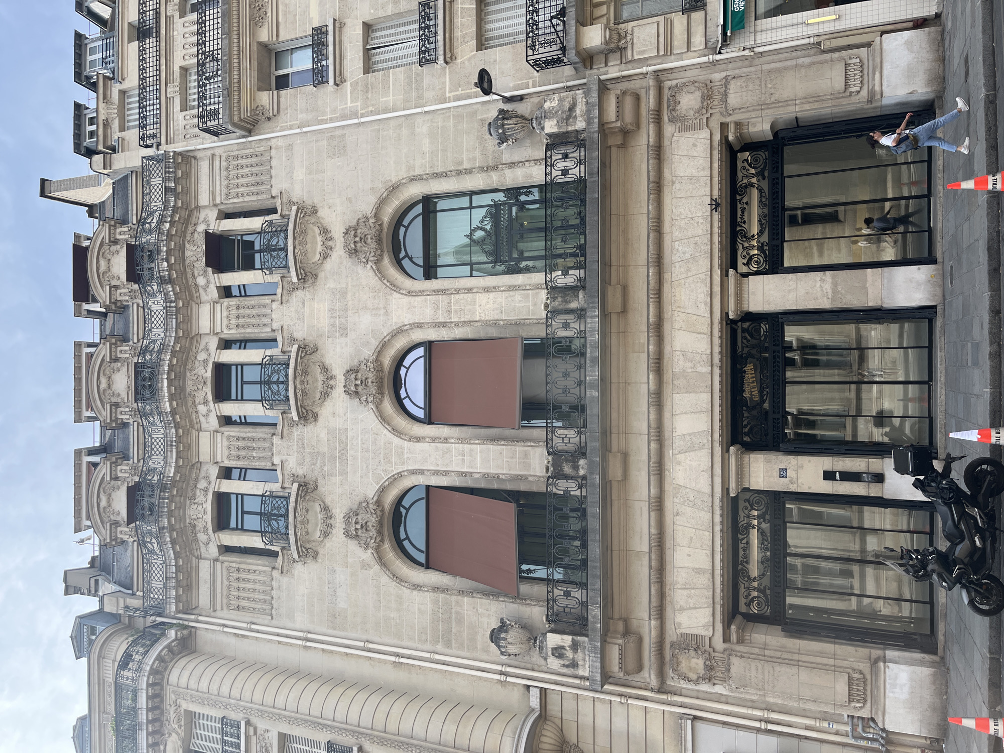 This image shows the exterior of a multi-story building in Paris, France, likely in a Haussmannian style, characterized by its stone facade, ornate architectural details, and wrought-iron balconies. The building features multiple arched windows on the upper floors, some adorned with brown awnings. The ground floor has large glass doors with black metal framing. In front of the building, a dark-colored motorcycle is parked on the sidewalk, and two orange traffic cones are visible. A woman wearing a white t-shirt, blue jeans, and a backpack is walking on the sidewalk. The sky above is overcast. Reflections in the ground floor glass reveal the interior of the building, showing some architectural features and at least one person walking inside. The facade is decorated with sculpted faces and elaborate moldings.