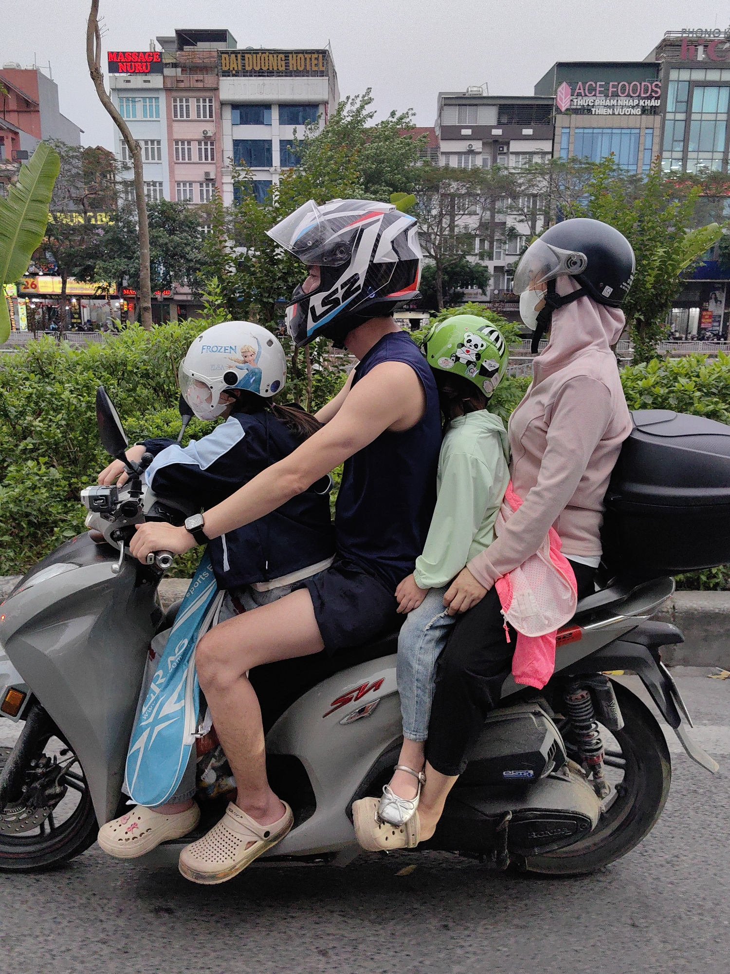 A family of four, including two children, ride on a scooter in Hà Nội, Vietnam. The adult male drives, while the adult female sits behind two small children. All are wearing helmets. The driver wears a sleeveless shirt and sandals; the young girl in front wears a "Frozen" helmet, sandals, and carries a bag with the words "Sports RAS." The second girl wears a green helmet with a panda bear. The street scene is visible, including buildings such as "ĐẠI DƯƠNG HOTEL" and "ACE FOODS THỰC PHẨM NHẬP KHẨU THIÊN VƯƠNG Q" visible in the background.