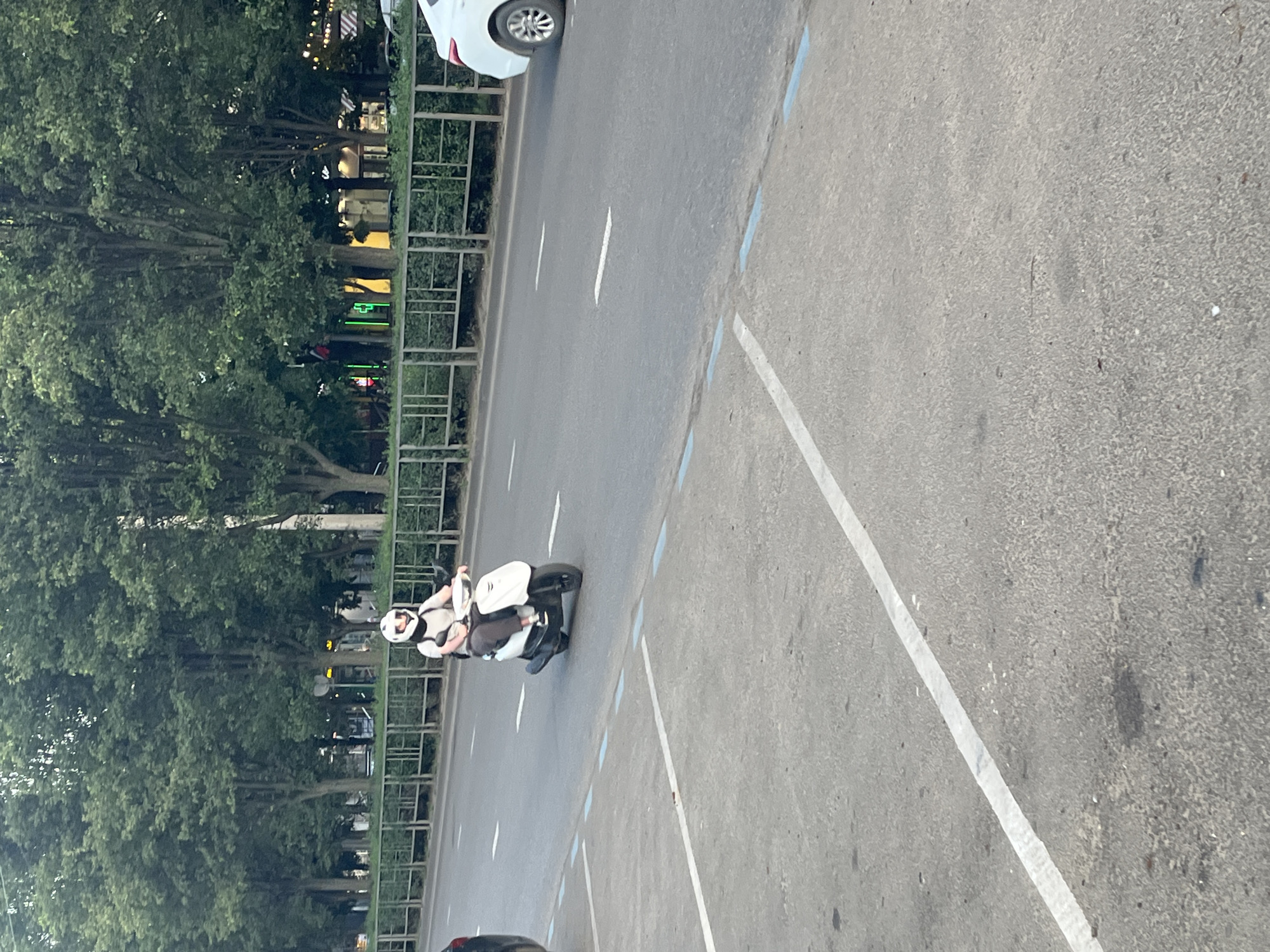 A person wearing a white helmet rides a white scooter on a paved road. The person is wearing a light-colored t-shirt and dark pants. The scooter has a dark seat and a dark rear storage compartment. The road has white lane markings. In the background, there are trees, a fence, and buildings with illuminated signage. A white car is visible on the right side of the road. The foreground shows a paved area with white and blue line markings, suggesting a parking lot. The scene appears to be in a city environment, possibly Krasnodar, Russia, given the street layout and vegetation.