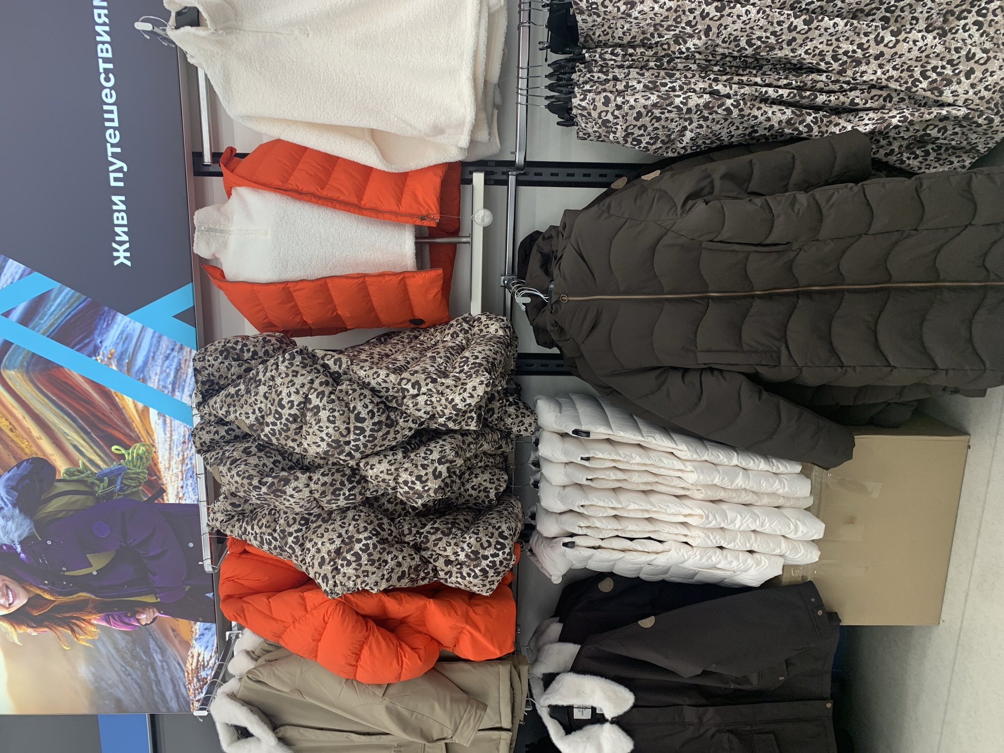 The image depicts a retail display of winter clothing. In the background, a large advertisement features a woman dressed in a purple winter coat and a backpack, with a mountainous, snowy landscape. Text in Cyrillic, reading "Живи путешествиями" (Live by travel), is displayed above the clothing racks.
The foreground showcases a variety of jackets and vests hanging on racks. From left to right, there is a beige jacket with a white fur collar, followed by a bright orange puffer vest, a patterned puffer jacket with a leopard-like print, and a white fleece jacket with a high collar. Below these, on another rack, are dark brown jackets, one with a prominent zipper and wavy quilting, and a row of neatly folded white puffer vests. To the far right, another leopard-patterned garment, possibly a skirt or dress, is displayed. A cardboard box is visible beneath the white vests.
The scene is consistent with the interior of a clothing store in Hrodna, Belarus, likely specializing in outerwear or sporting apparel. There are no people visible in the image. The lighting is artificial, typical of an indoor retail environment.