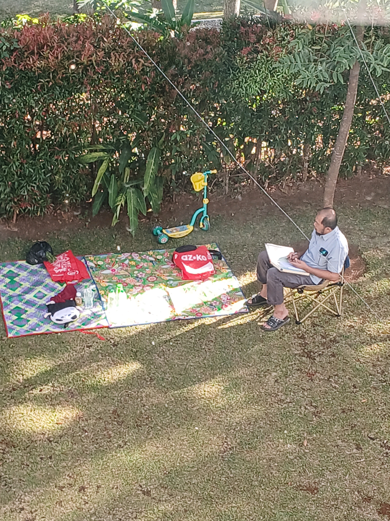 A man is sitting on a folding chair in a grassy area near a dense hedge of red and green foliage. The man is bald with a beard and is wearing a light blue short-sleeved collared shirt and dark gray pants. He is holding a white laptop on his lap and appears to be looking at it. In front of him on the grass, there are two picnic mats, one with a geometric pattern of purple, green, and white, and another with a floral pattern in green, pink, and yellow. On the first mat are a black garbage bag, a red bag with white lettering, and a maroon item. On the second mat, there are several clear plastic bottles and glasses, a red bag with "AZ-KO" written on it, and a white pillow. To the left of the mats, a yellow and teal scooter is propped up on its kickstand. The scene is outdoors, with dappled sunlight on the grass. The background includes trees and what appears to be a building or structure in the distance. The location is Arjasari, Indonesia.