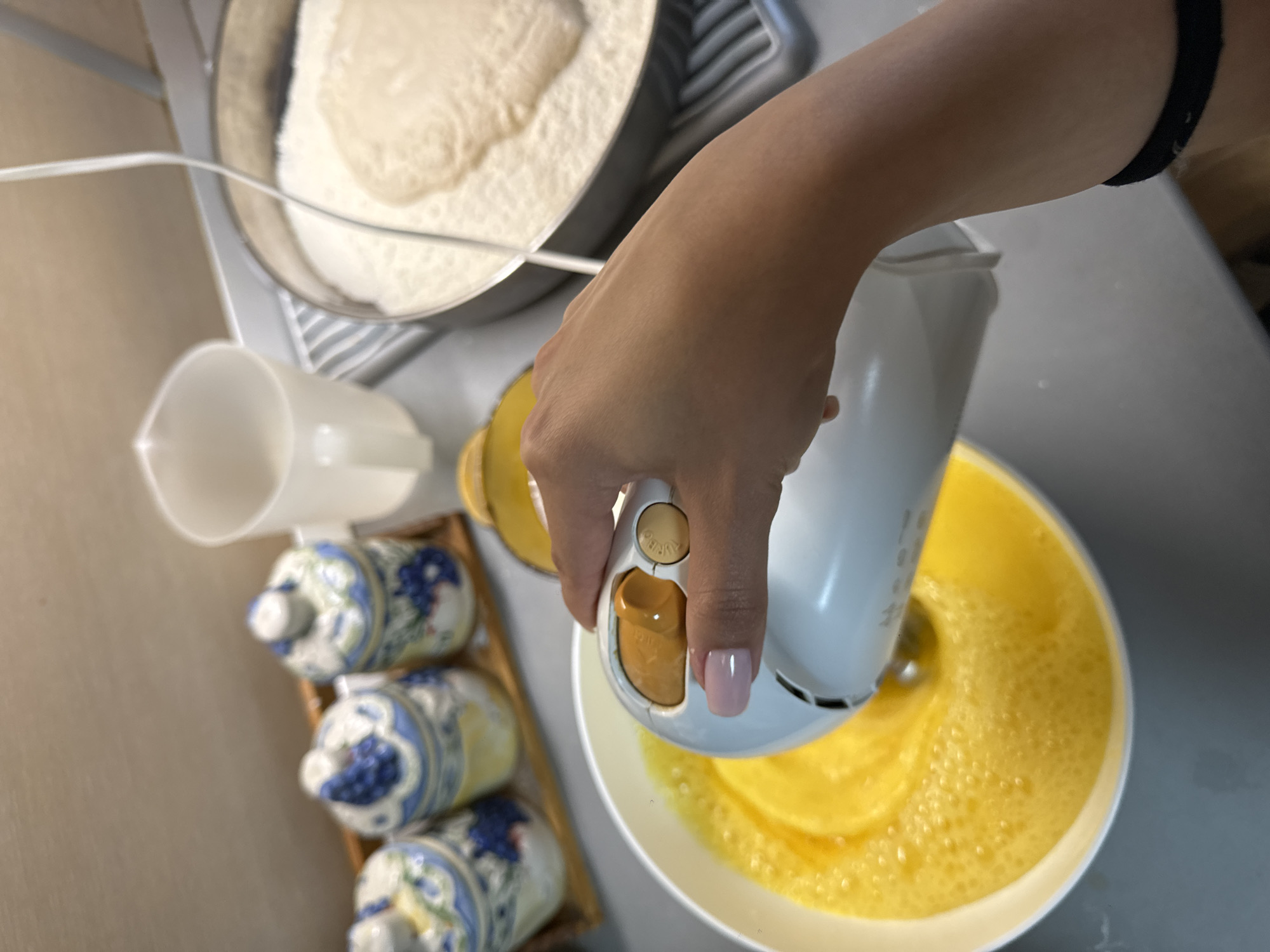 A person is using a handheld electric mixer to mix yellow liquid ingredients in a white bowl. The mixer is white with a yellow button. To the left, there is a container of white flour and a measuring cup. Below that, there are three decorative ceramic containers. The setting appears to be a kitchen. The image was not taken in Dupnitsa, Bulgaria.