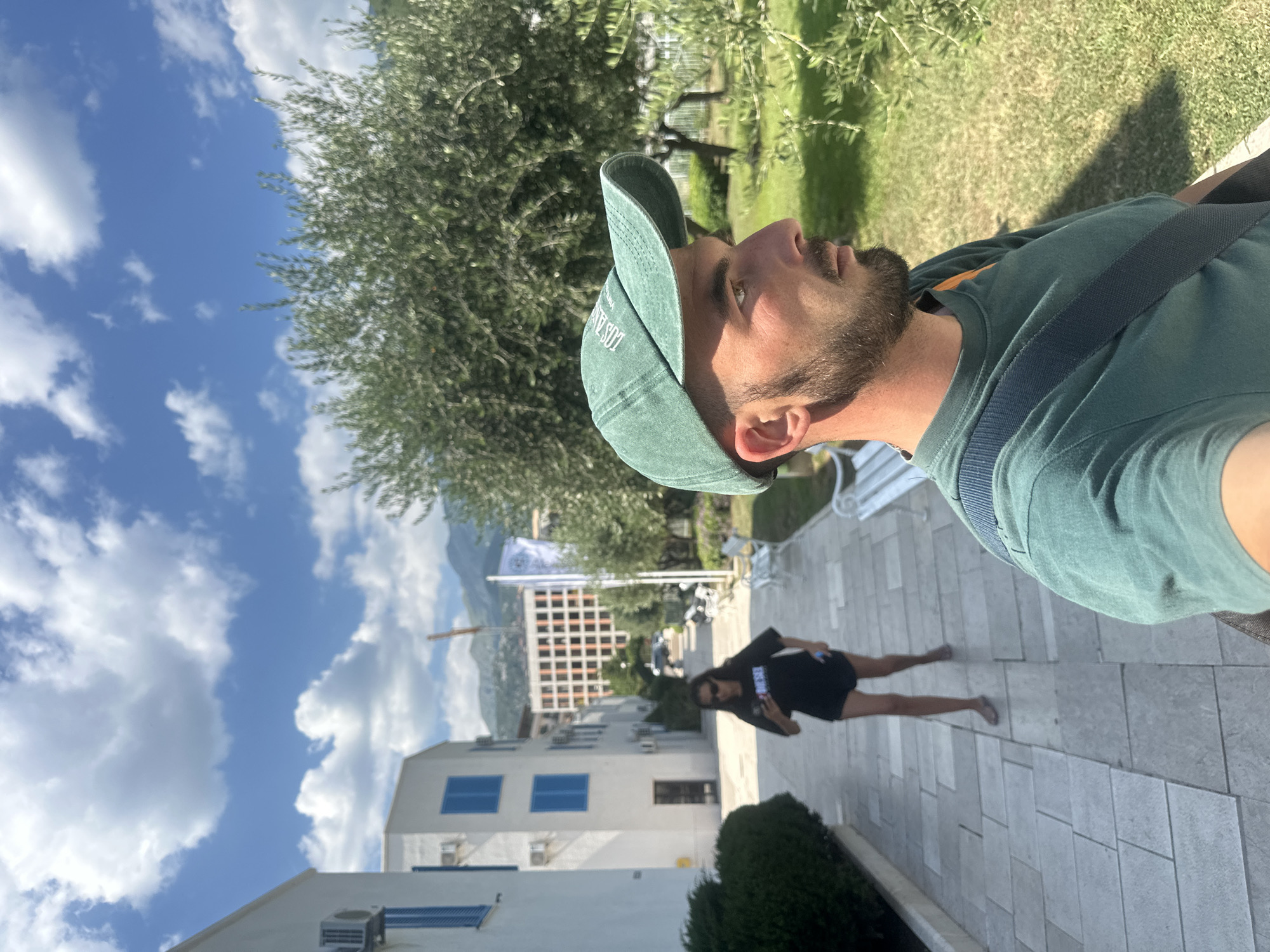The media file depicts a selfie taken by a man wearing a green t-shirt and a green baseball cap. He has a beard and is looking up and to the right. A woman in a black dress and sunglasses is visible in the background, walking away from the camera and holding a phone. The scene appears to be outdoors, with a paved walkway, grass, trees, and buildings. In the distance, there are mountains and a large building with a grid-like facade. A white flag on a pole is also visible. The sky is blue with scattered white clouds, suggesting it is daytime with fair weather. The man's expression is neutral. There is text visible on the man's cap, which appears to read "LOS A..." and on the woman's dress, which is not clearly legible. The setting is likely a public area or resort in Budva, Montenegro, given the context.