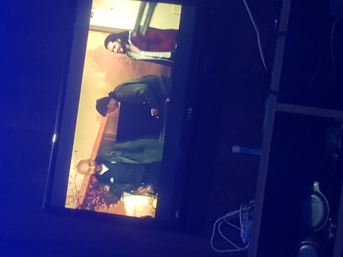 A television displays a scene with three figures. On the bottom left, a bald man in a dark suit smiles. Above him, a man in dark clothing bends over. At the top right of the screen, another man with long hair wears a red and black plaid shirt over a light-colored jacket. The background has a reddish-brown hue with what appears to be a pattern or design. The television sits on a dark wooden shelf where cables and headphones can be seen. The lighting is dim.
