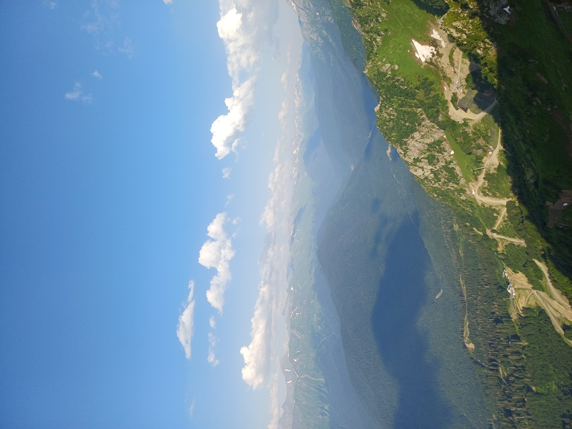The image depicts a vast mountainous landscape under a clear blue sky with scattered white clouds. In the foreground, a steep, green mountainside is visible, with patches of snow and rocky outcrops. A winding dirt road snakes its way up the mountain, with some structures, possibly ski lifts or buildings, situated along it. The middle ground features rolling, tree-covered hills and valleys, with shadows cast by clouds creating a textured effect. In the background, a range of distant, hazy mountains can be seen, some with snow-capped peaks. The overall impression is of a scenic, possibly resort-like mountain environment in Estosadok, Russia. There are no people or animals visible in the image.