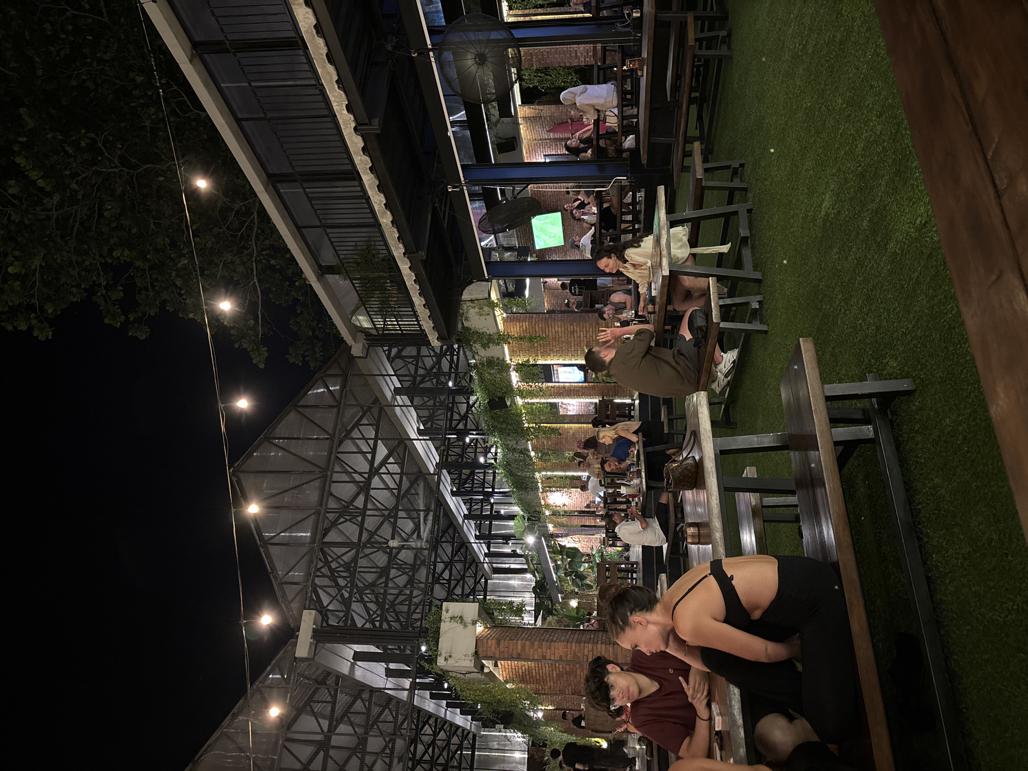The image depicts an outdoor restaurant or bar at night in Kuta Selatan, Indonesia. The venue features multiple levels with metal framework, brick pillars adorned with greenery, and string lights. Several patrons are seated at tables, engaging in conversation and eating. A couple is in the foreground, with one person bending toward the other. The establishment's flooring is artificial grass. A television screen is visible in the middle ground, and ceiling fans are present.
