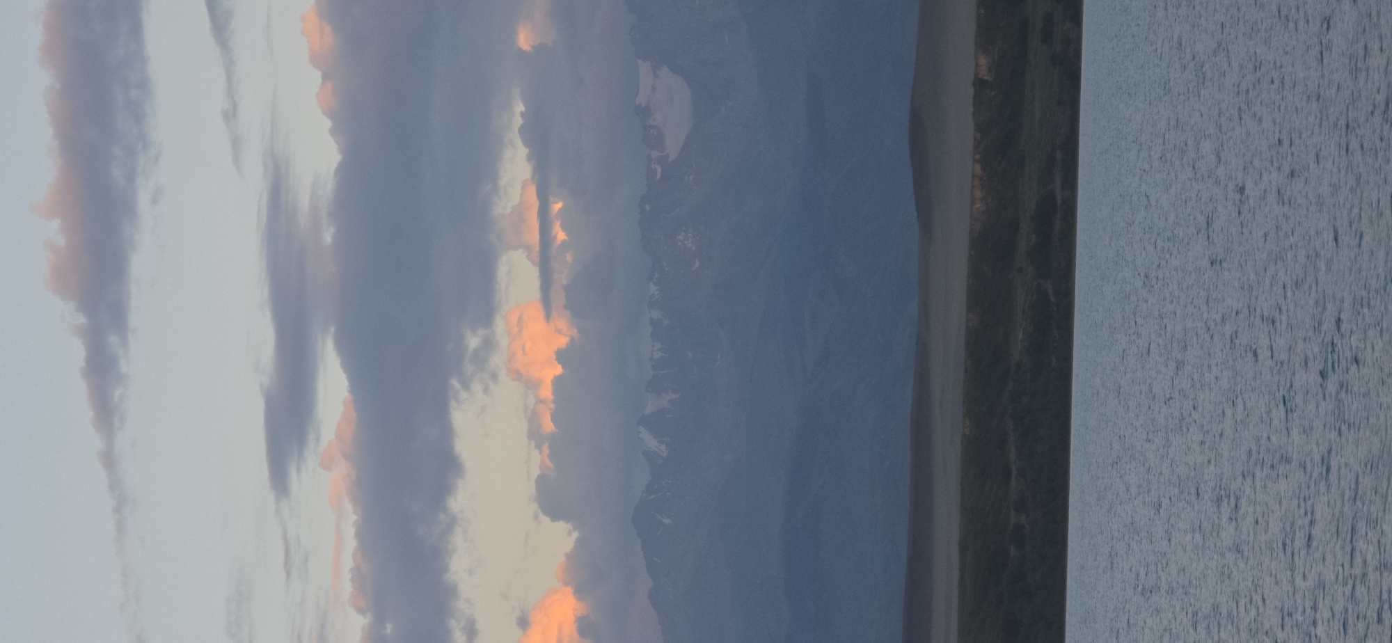 The image depicts a serene landscape with a body of water in the foreground, reflecting the soft hues of the sky. In the mid-ground, a gently sloping hill is visible, leading towards a majestic mountain range in the distance. The mountains are capped with snow, adding a dramatic contrast to the earthy tones of the terrain. The sky is adorned with a scattering of clouds, some tinged with the warm glow of sunset or sunrise, while others are a soft, muted gray. The overall scene evokes a sense of tranquility and natural beauty. The location is identified as Pristan'-Przheval'sk, Kyrgyzstan, suggesting a setting within this region. No people or specific man-made structures are visible in the image, focusing entirely on the natural elements.