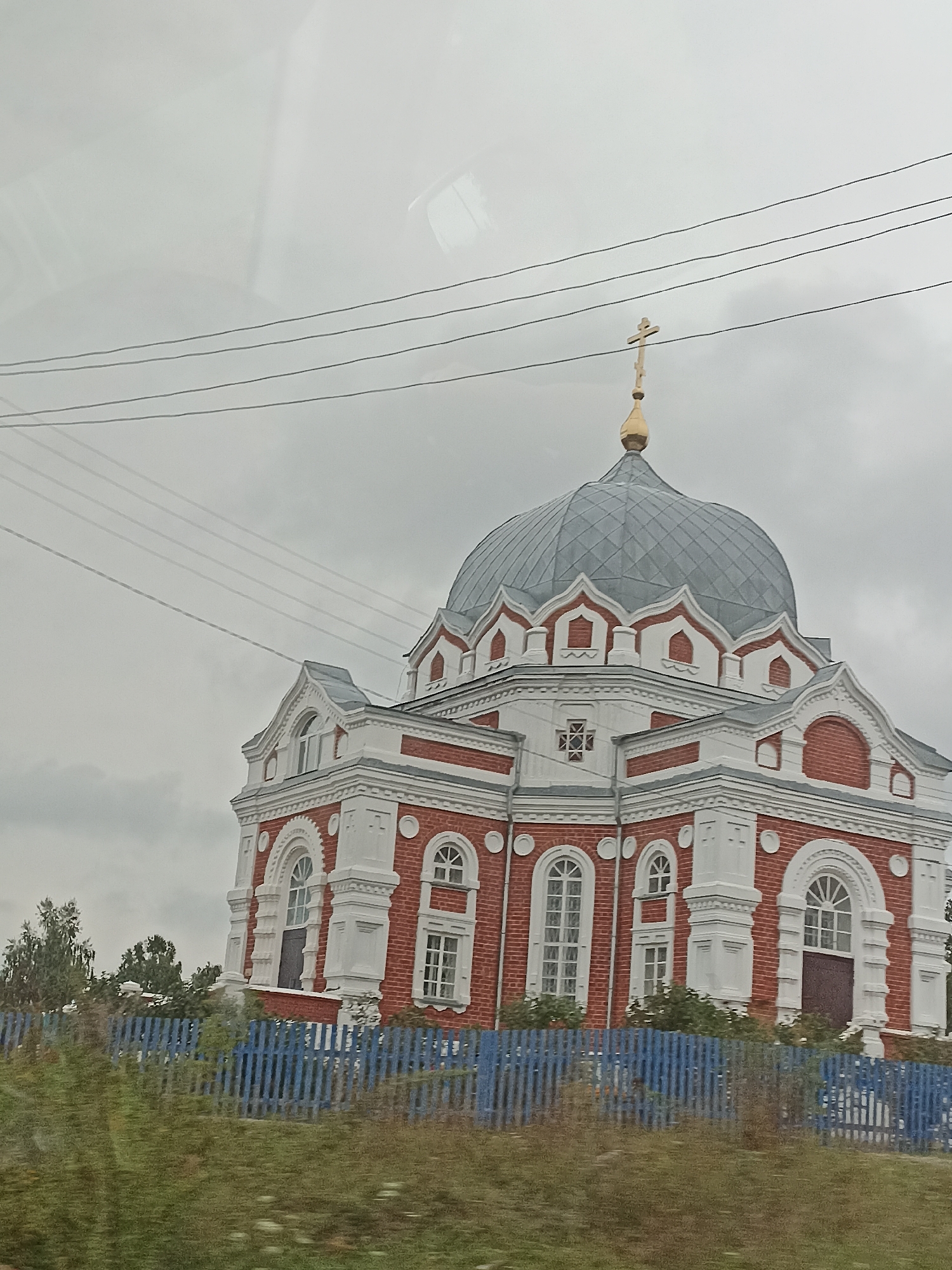 The image shows an Eastern Orthodox church with a red brick facade and white decorative elements, topped with a gray dome and a golden cross. The church is situated behind a bright blue wooden fence. In the foreground, there is overgrown grass and foliage. Overhead, multiple power lines are visible against a cloudy sky. The scene is likely located in the village of Zavyalovo, Russia, given the architectural style which is typical for rural Russian churches. No people are present in the image.