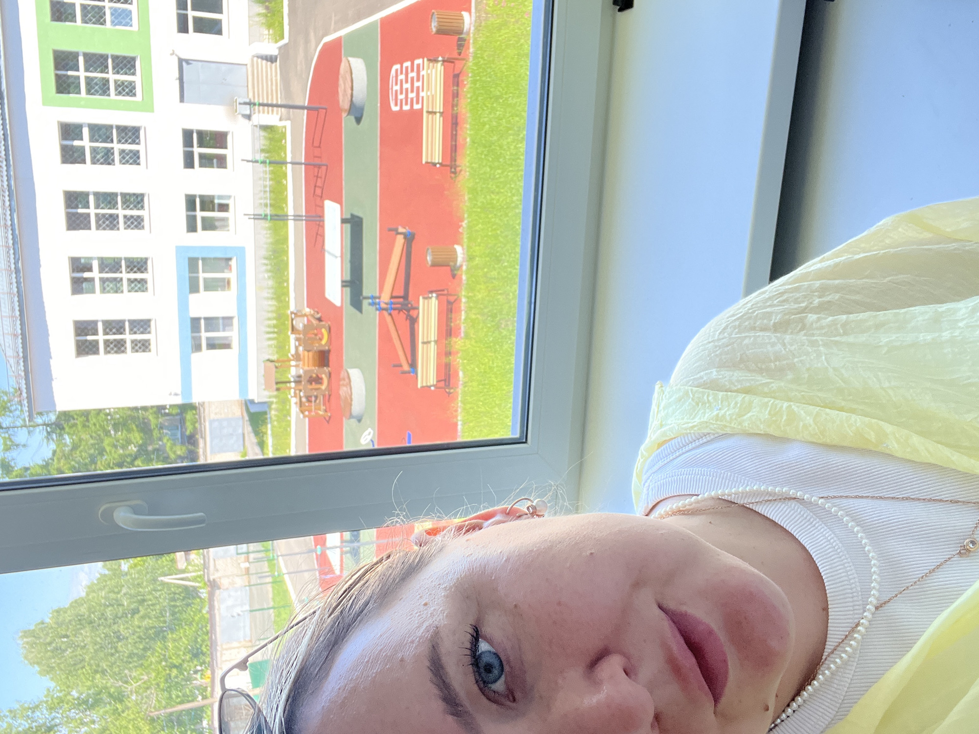 A woman is taking a selfie through a window, with a playground and a building visible in the background. The woman's face and upper torso are in the foreground, slightly to the left of the frame. She has light skin, blue eyes, and light brown hair. She is wearing a white tank top, a yellow open-front garment, and two necklaces, one with small pearls and another with a pendant. She also has a small hoop earring with a pearl in her visible ear.
The background shows a brightly lit outdoor area with a red and green-colored playground surface. There are several playground structures, including benches, trash cans, a small table, and what appears to be a climbing structure. Beyond the playground, a multi-story building with white walls and colorful accents (blue, green, and white) is visible. The building has multiple windows with gridded panes. Trees and a fence are also seen in the distance. The overall scene suggests a sunny day. This image was taken in Chaikovsky, Russia.