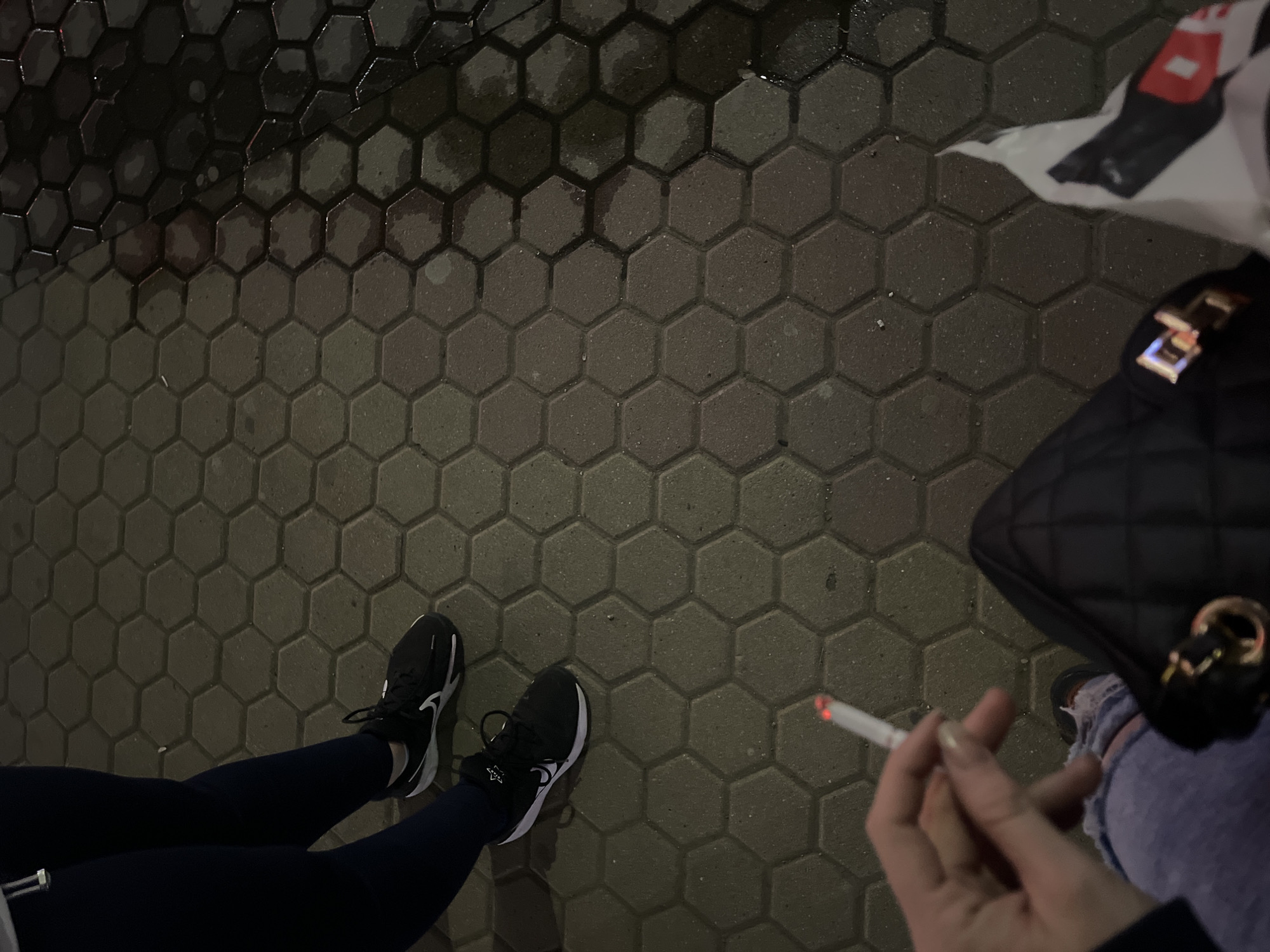 The image shows a person holding a lit cigarette on a paved surface in what appears to be an urban environment. The pavement is comprised of grey, hexagonal blocks. The lower portion of a person is visible, wearing dark-colored leggings and black sneakers with white accents. To the upper-right is a black, quilted handbag and part of a white plastic bag. The image gives no specific indication of being in Sofia, Bulgaria.