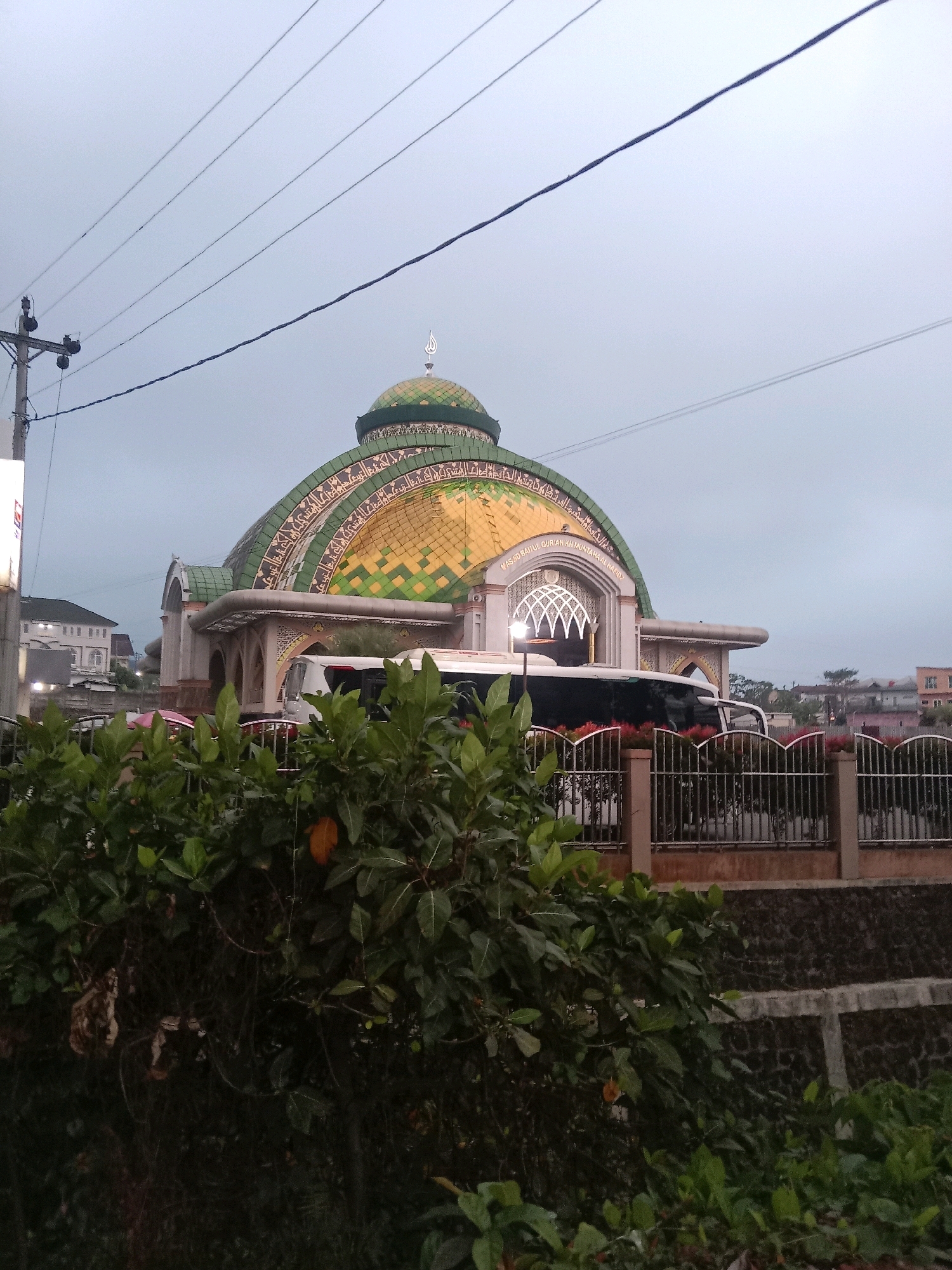The image depicts a mosque with a large, green and yellow domed roof in Mojotengah, Indonesia. The mosque features ornate architectural details, including Arabic calligraphy around the dome and a decorative archway above the entrance. Several buses are visible parked in front of the mosque, suggesting it is a place of worship and possibly a travel hub. Lush green foliage in the foreground partially obscures the view of the mosque and buses. The sky is overcast, indicating a cloudy or possibly rainy day. Electrical wires are strung across the top of the image, common in urban or semi-urban environments. The overall scene captures a moment of quietude in a public space, with a focus on the architectural grandeur of the mosque.