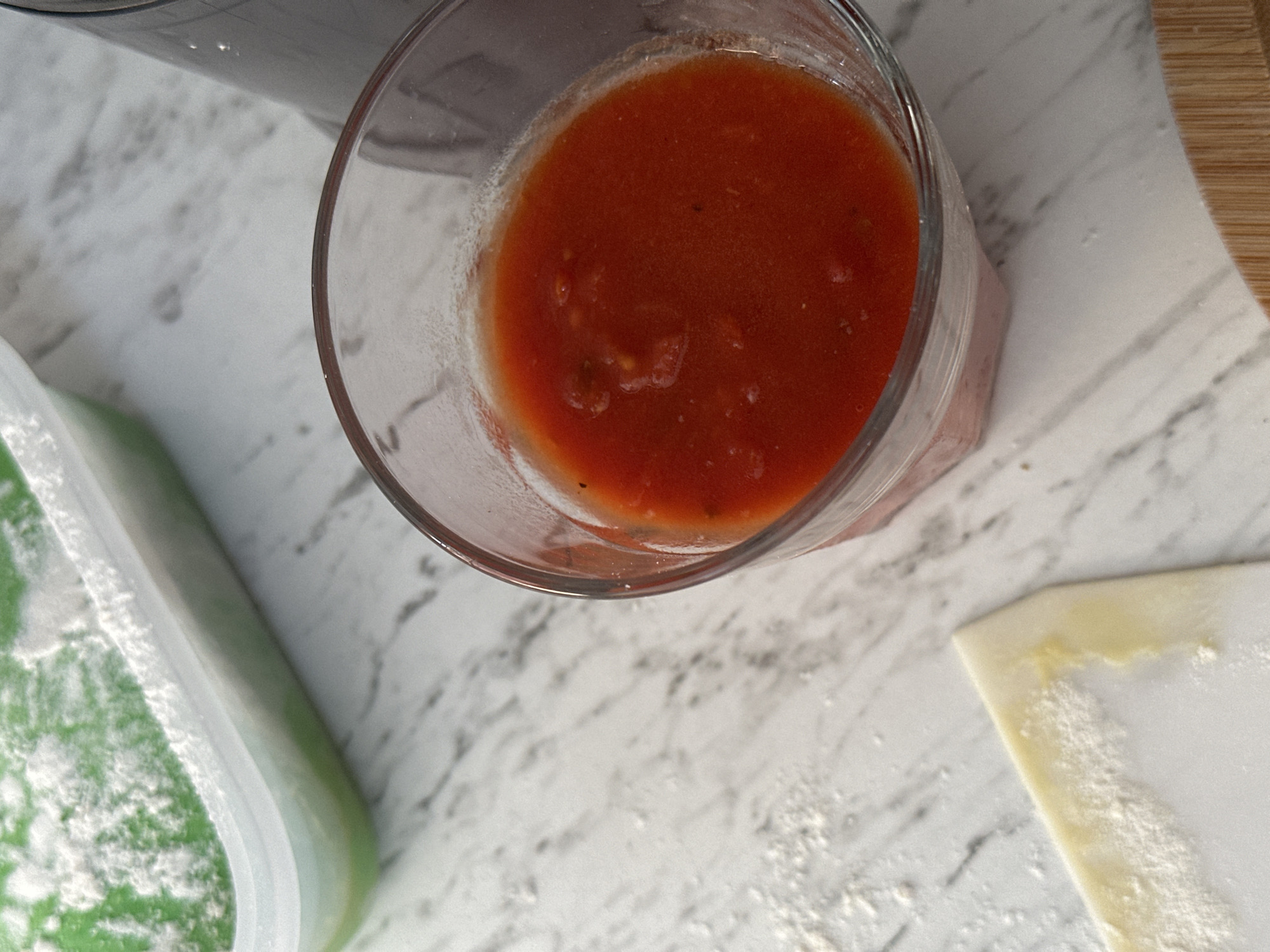 A close-up, high-angle shot shows a glass of red sauce and a plastic container with a green substance dusted with white powder, both on a marble-patterned countertop. A small piece of dough, dusted with flour, is visible in the lower left corner. A blurred dark object, possibly a blender, is in the background on the right. The scene appears to be in a kitchen. The image does not contain information to locate it in Moortown, Ireland.