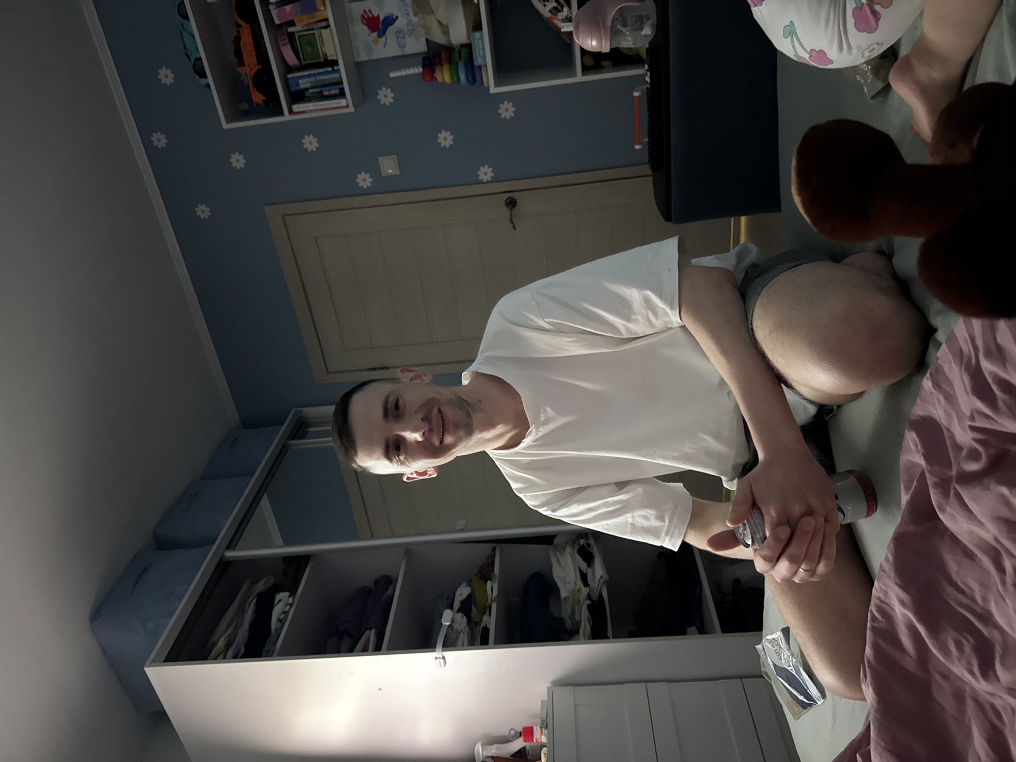 The image captures a man smiling directly at the camera, seated on a bed or sofa in what appears to be a child's bedroom. The man, in his late 20s to early 40s, has short dark hair and stubble, and is wearing a white t-shirt and dark shorts. He holds a silver and red beverage can, partially revealing the text "HOPS". A simple band is visible on his left ring finger.
To his right, partially visible on the bed, is a pink duvet, a brown stuffed animal, and a leg wearing a white diaper with pink and green floral patterns, suggesting the presence of a baby or toddler. Near his right leg, a small clear package with the Cyrillic text "КЕМ" (KEM) is visible.
The room features a light blue wall adorned with white daisy decals. A light-colored wooden door with a dark handle is visible in the background. Above the door, a white wall-mounted shelf unit holds various items, including books (one with Cyrillic text "БАКЕРИ" - BAKERY), and children's toys like a colorful stacking ring and a pink pencil sharpener. To the man's left, a white wardrobe or storage unit with multiple compartments, some containing clothes, stands against the wall. Three light blue fabric storage boxes are stacked on top of this unit.
The lighting is soft, likely artificial or indirect, contributing to a casual and relaxed atmosphere. The man's wide smile conveys happiness and engagement. The setting suggests a home environment, possibly a family living space in Moscow, Russia.