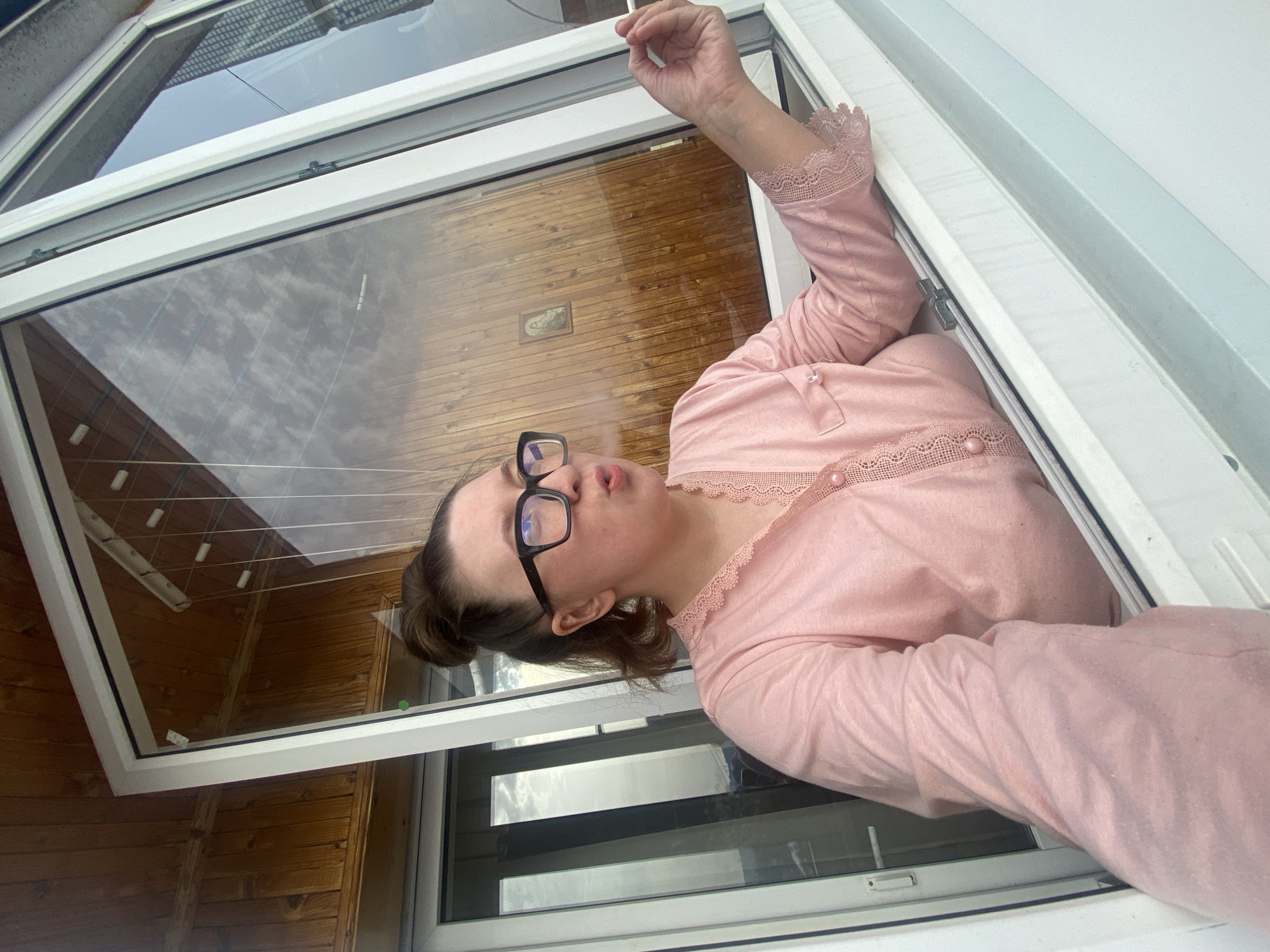 A woman with brown hair tied in a bun and wearing black-framed eyeglasses is observed leaning out of an open, white-framed window, exhaling smoke from a cigarette. She is wearing a pink long-sleeved top featuring lace trim at the neckline and cuffs. Her right hand holds the cigarette. Behind her, visible through reflection in the window, is light-colored wooden paneling with a small framed religious icon depicting a saint. Additional reflections in the window display an urban environment, including a multistory building and a partially visible clothes drying rack with white pegs. The scene is located in Chaikovsky, Russia.
