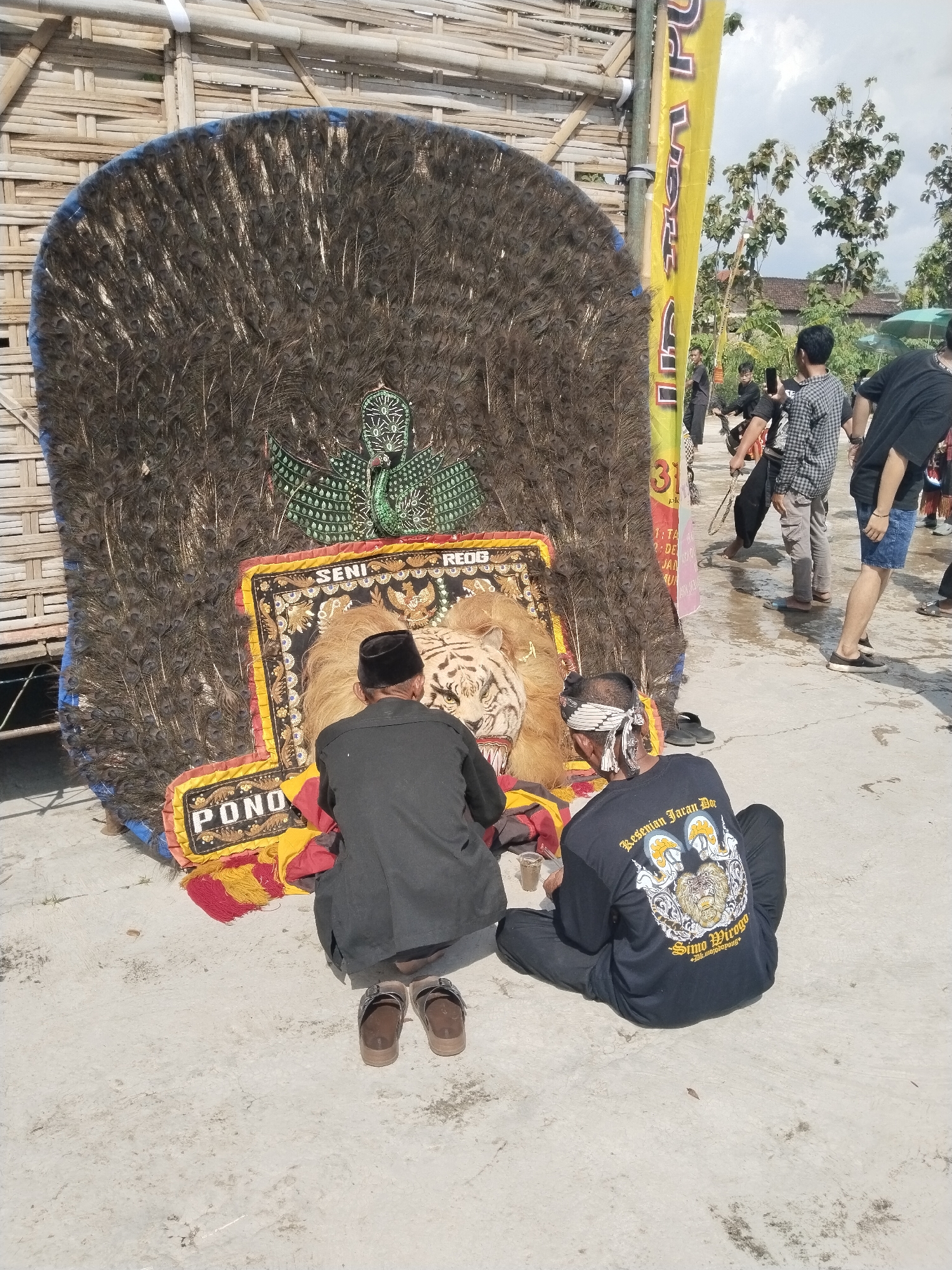 The image shows two men crouching in front of a large traditional Ponorogo tiger dance mask decorated with peacock feathers, with the words "PONO," "SENI REOG," "Besenian Jaran Dor," and "Simo Mirogo" printed on it, suggesting the scene is in Mojodoyong, Indonesia. Other people are standing in the background. The ground appears to be a paved surface.