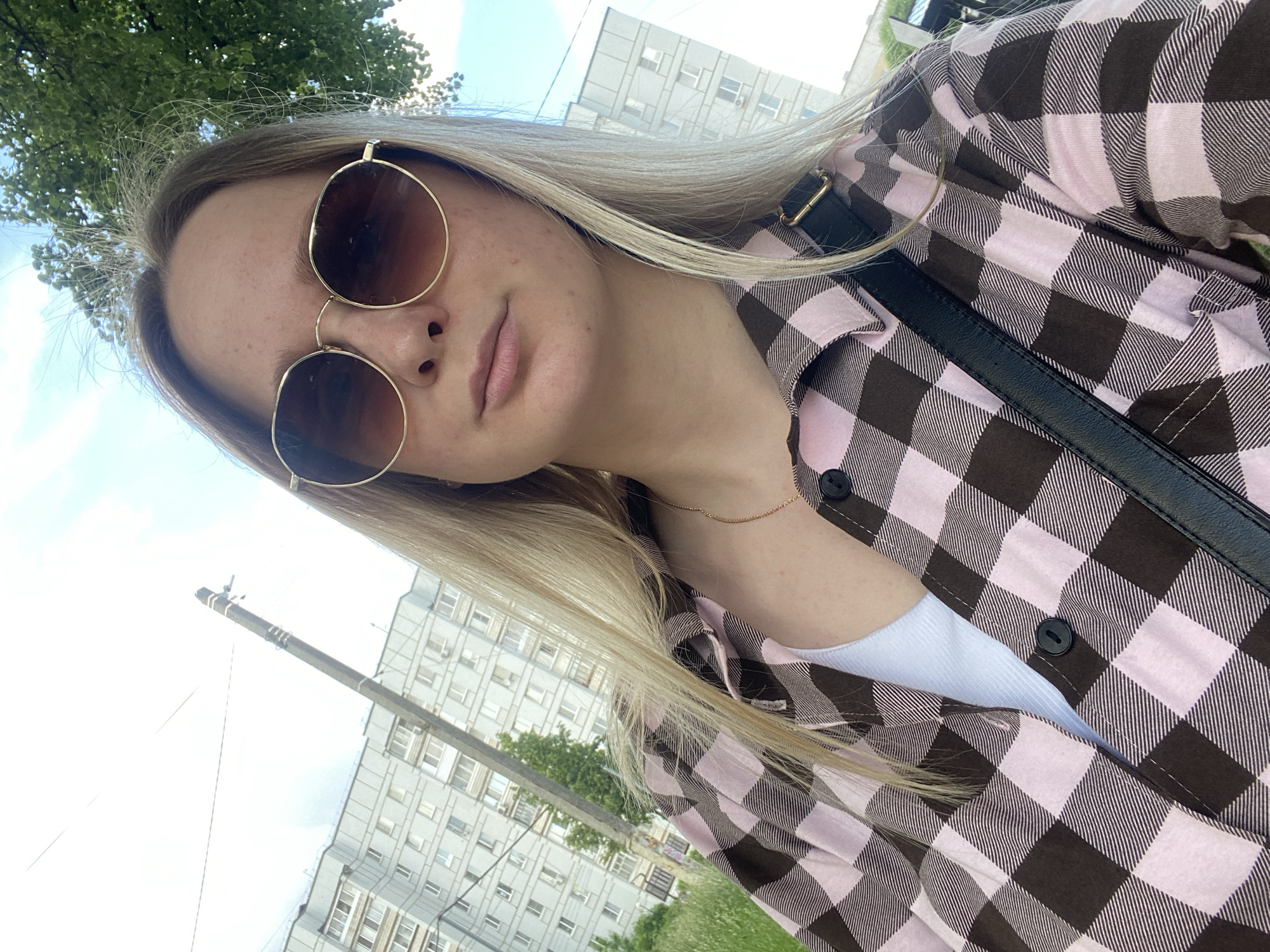 In Chaikovsky, Russia, a young Caucasian woman with blonde hair and sunglasses takes a selfie. She is wearing a pink and brown checkered shirt over a white top and a black strap is visible across her chest. In the background, a multi-story apartment building, a utility pole, and trees are visible under a partly cloudy sky.
