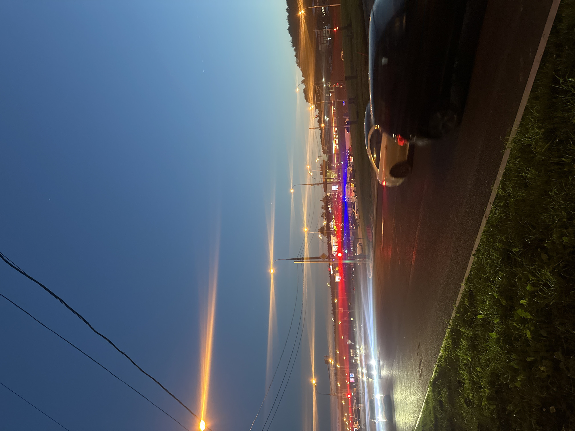 The image depicts a street scene in Moscow, Russia, during dusk or early night. The sky is a deep blue with streaks of orange and yellow from streetlights and possibly the setting sun. Several streetlights illuminate the scene with warm, bright glows. Power lines are visible against the sky.
The road in the foreground appears wet, possibly from recent rain, and reflects the artificial lights. Several cars are present, with some showing motion blur due to the low light conditions and likely long exposure photography. A silver car is visible in the middle ground, and a black car is in the foreground, partially obscuring the view. In the distance, there are more lights from buildings and traffic, creating a vibrant, somewhat blurred urban landscape. The background features a treeline and the architecture of commercial buildings. The overall atmosphere is that of a busy urban environment at twilight, with the artificial lights beginning to dominate the natural light.