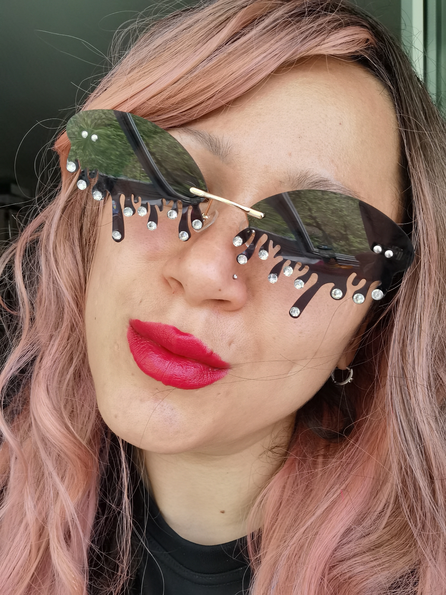 A person with pink hair, wearing novelty sunglasses with black "dripping" edges and small gemstone embellishments, is facing the camera. The person is wearing red lipstick and has a piercing in their left nostril. An earring is visible in the left ear. The background is blurred, suggesting an outdoor setting with a glimpse of green foliage reflected in the sunglasses. There is no indication of the location being in the city of Chaikovsky, Russia.