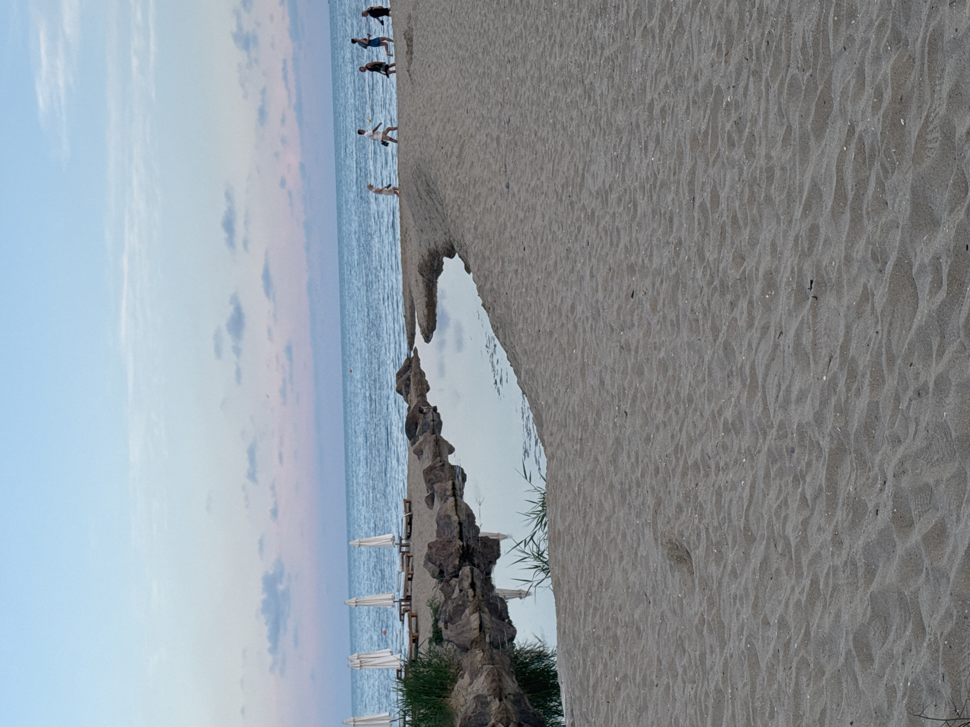 A wide, sandy beach slopes gently towards the ocean under a pastel-colored sky, with soft clouds in shades of pink and blue. In the mid-ground, a small, calm body of water, likely a tidal pool or lagoon, is nestled among dark, rough rocks and sparse green reeds. Several white beach umbrellas are set up in the background on the sand, accompanied by wooden lounge chairs. A group of five individuals are walking along the shoreline near the water's edge. On the left, the umbrellas are lined up, and on the right, the people are spread out, some walking in unison, others more spaced apart, as they move away from the viewer. The sand in the foreground is textured with small ripples and scattered debris. The overall scene captures a tranquil beach at dusk.