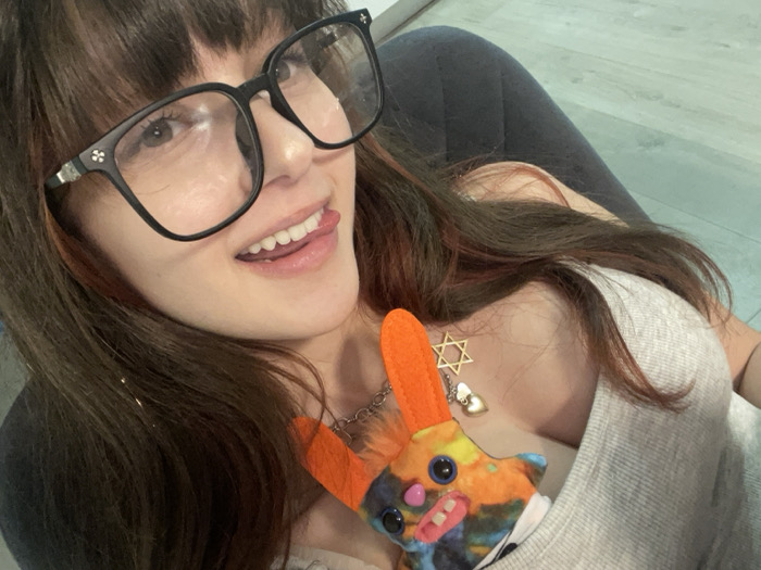 The image displays a close-up, high-angle shot of a female subject in an interior setting in Raanana, Israel. The specific address or type of room within the city cannot be determined from the visual information. The subject, partially visible from the chest up, has long, dark brown hair with reddish highlights and is wearing large, black-framed eyeglasses. She is looking towards the camera with her mouth slightly open and her tongue extended. She wears a light gray, ribbed or knit top. Around her neck is a silver-toned chain necklace featuring a golden Star of David pendant and an additional small, white, heart-shaped charm. A multi-colored stuffed toy, characterized by prominent orange ears, cartoonish eyes, and visible white teeth, is tucked into her top near her chest. The toy displays abstract patterns in blue, orange, yellow, pink, and black hues. The background consists of a dark, possibly upholstered, surface on the left and a light-colored floor visible in the upper right.