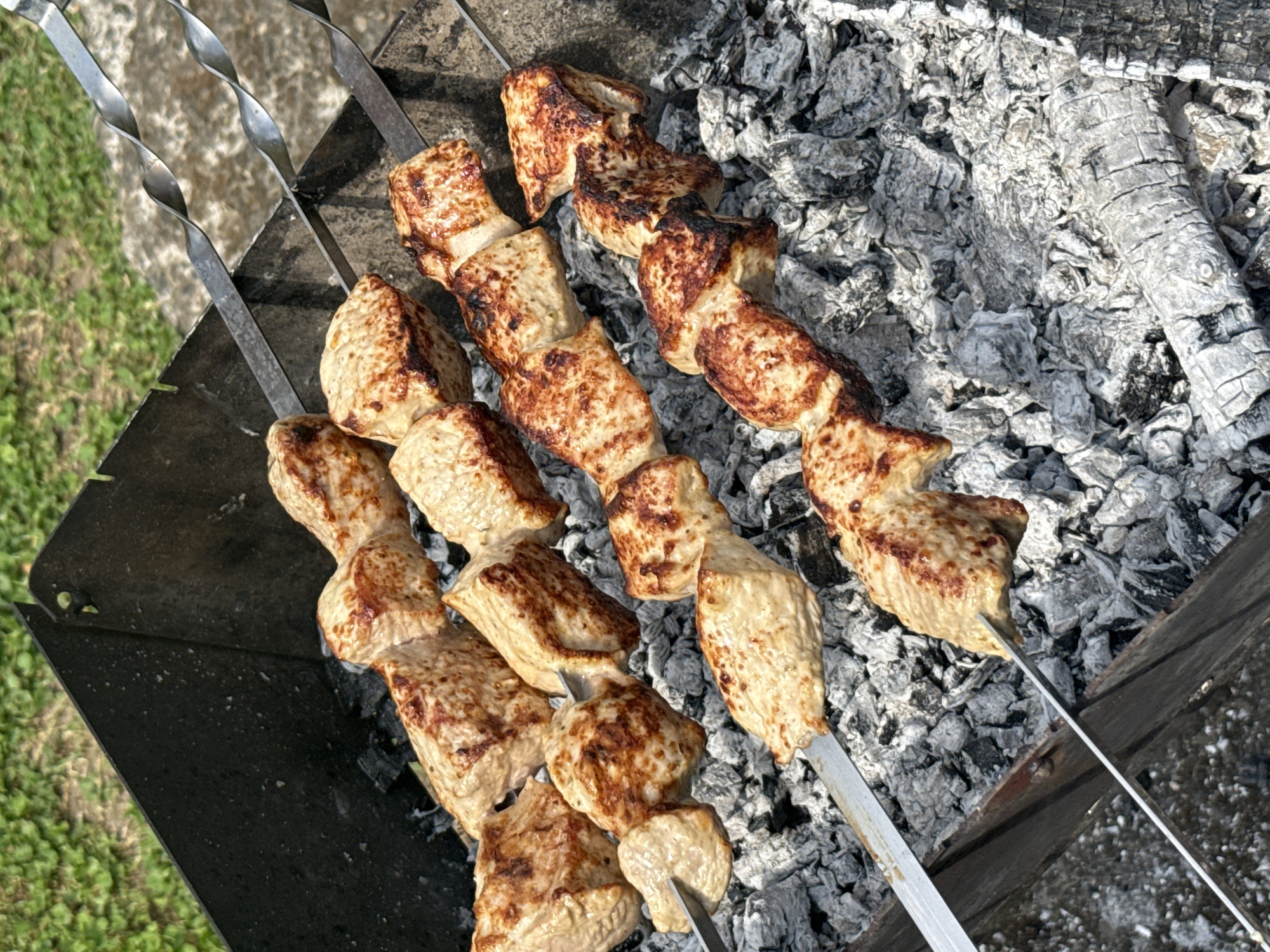 Four metal skewers, each loaded with pieces of raw, marinated meat, are positioned horizontally over a bed of white and grey ash in a metal barbecue grill. The meat is a pale pinkish-white and appears to be chicken or pork. The grill is made of dark metal, with a slightly rusted appearance on the edges. The background is out of focus, showing green grass and a rough, grey surface that might be concrete. The lighting suggests an outdoor setting, likely during the daytime. There are no people or other discernible objects in the scene.