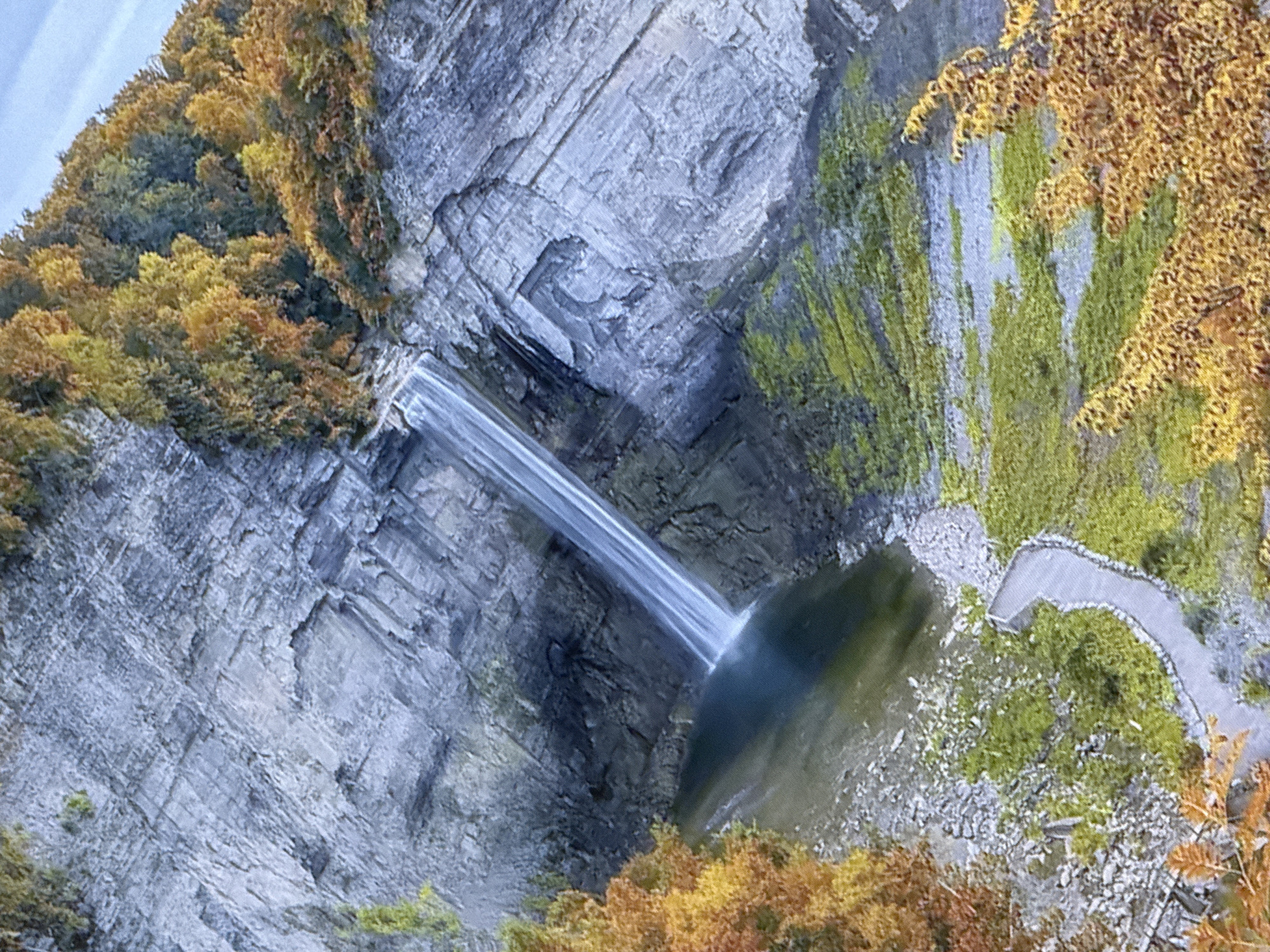 The image depicts a tall, narrow waterfall cascading down a sheer rock face into a dark pool of water. The rock face is layered and appears to be composed of gray stone with some patches of green moss. Surrounding the waterfall and pool, the landscape is covered in autumn foliage, with trees displaying vibrant shades of orange, yellow, and green. A winding pathway with a stone railing is visible, leading away from the base of the waterfall and through the vegetated area. The sky above is a pale blue with some wispy clouds. The scene is captured from a high vantage point, looking down onto the waterfall and its surroundings. The specific location in the city of Simferopol, Ukraine is not discernible from the image itself.