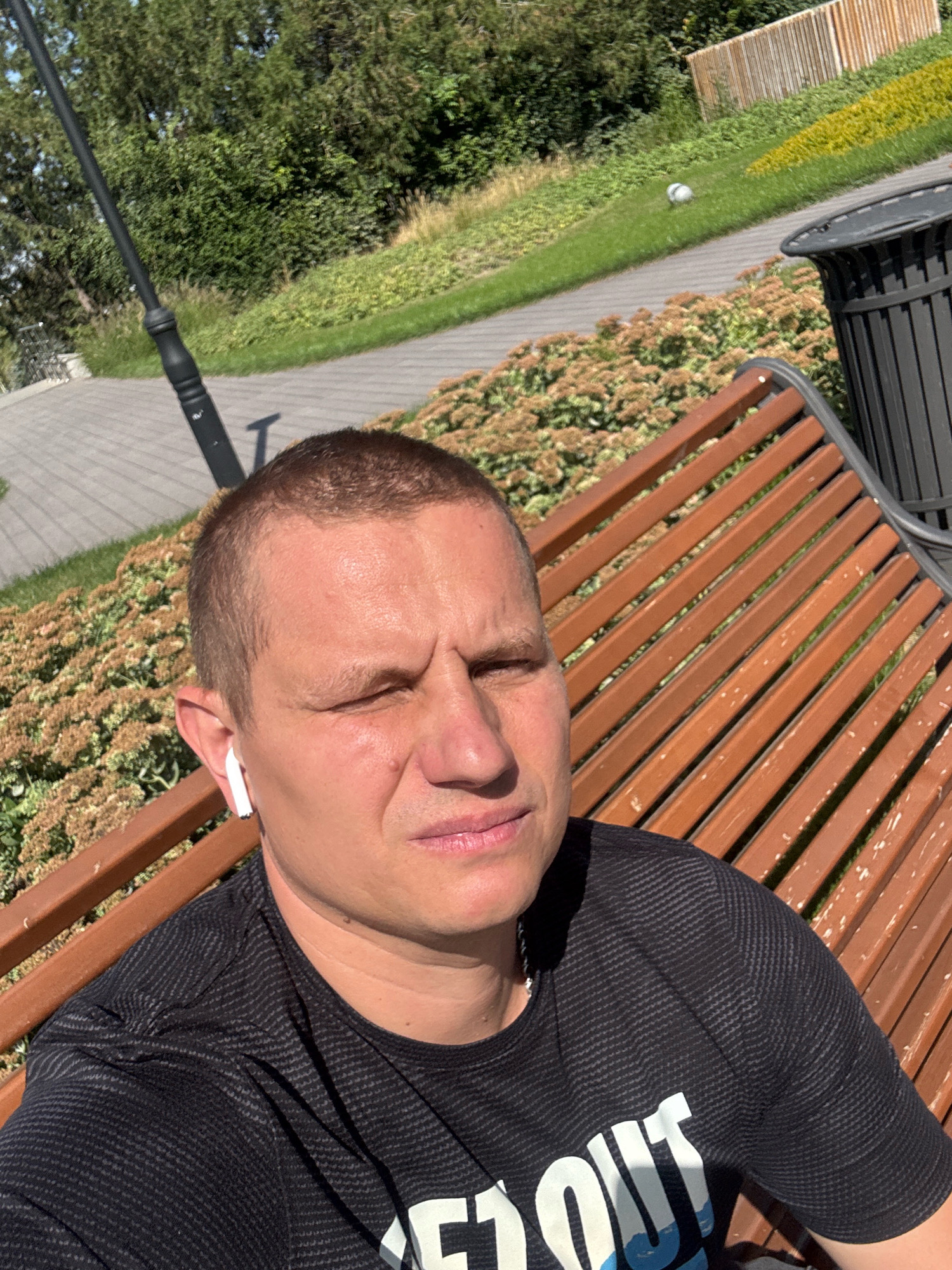 A man with a buzz cut and a light beard is sitting on a park bench, looking to the right of the camera. He is wearing a black t-shirt with a light-colored graphic design. He has white wireless earbuds in his ears. The bench is made of wooden slats and is in a park setting. Behind him, there are bushes with small, light-brown flowers and a paved walkway. In the background, there are trees, a lamppost, and a black trash bin. The scene appears to be in daylight, under a clear sky, suggesting it is a sunny day. The location is the Almaty Central Park in Almaty, Kazakhstan.