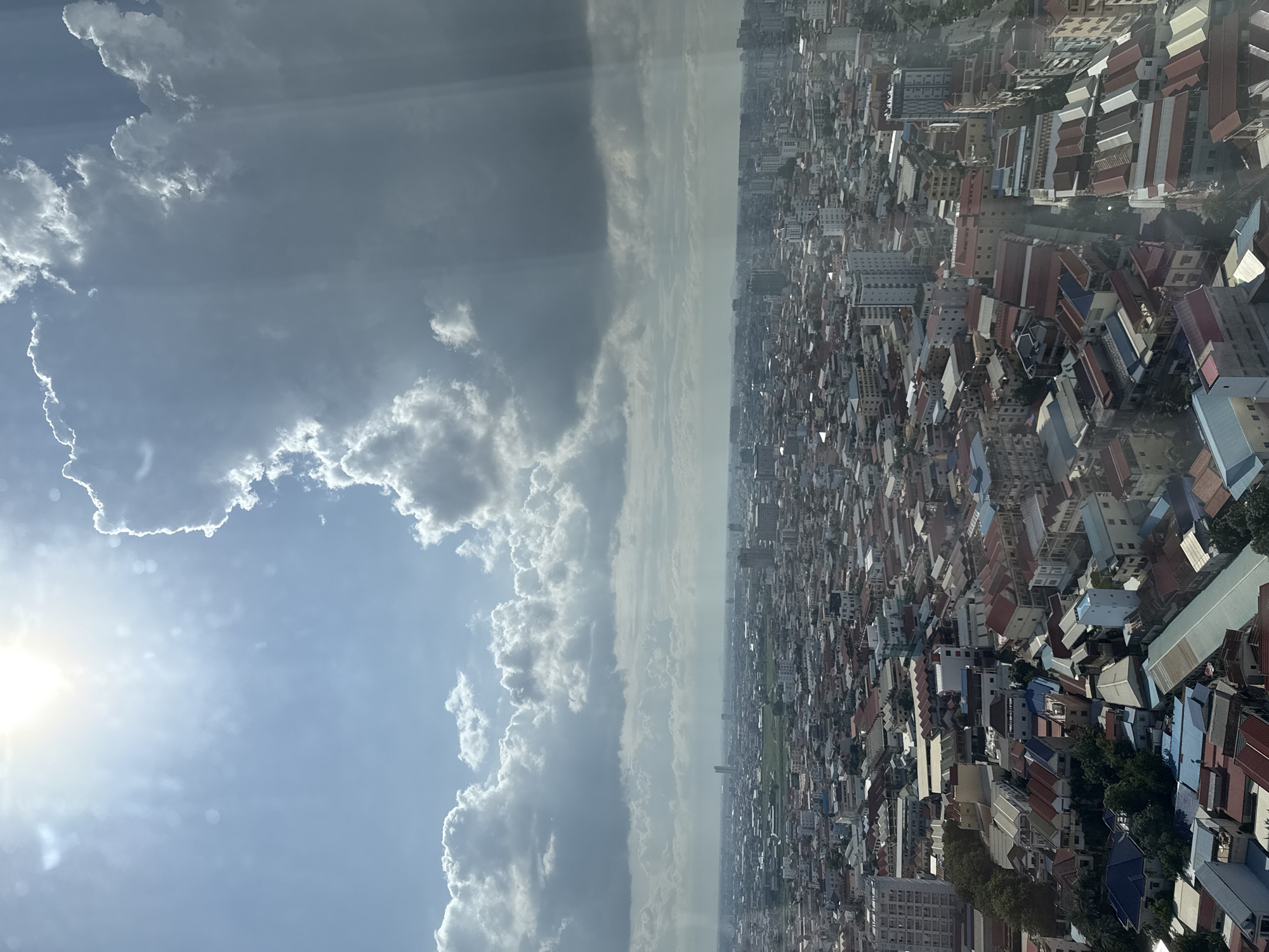 An aerial view looks down on a densely packed urban landscape, likely within Khan Mean Chey, Cambodia. The city stretches out to the horizon, a mosaic of buildings with varied roof colors, including red, gray, and blue, and some with corrugated metal. Tall buildings, some appearing to be modern high-rises, are interspersed with lower-rise structures. Vegetation, in the form of trees, is visible in pockets throughout the city. The sky above is a dramatic mix of bright sunlight and heavy, dark clouds, with visible rays of light breaking through. The overall impression is one of a sprawling, active city under a dynamic sky.