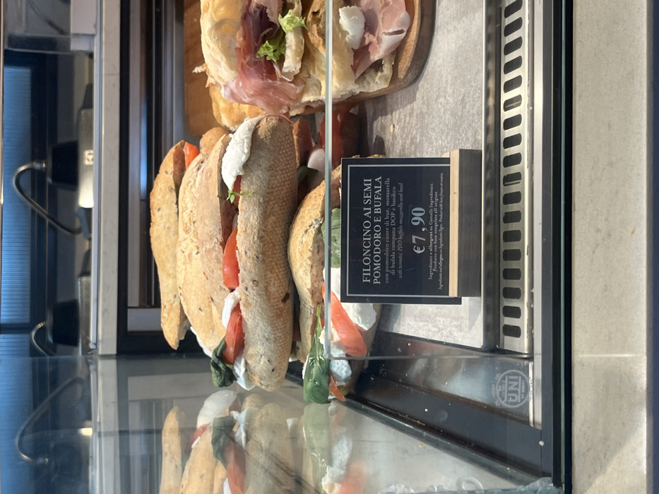 The image shows a well-lit food display case, likely in a deli, cafe, or bakery. The perspective is from slightly above, looking into the glass counter where various sandwiches are presented for sale. The overall scene suggests a commercial food establishment.
On the upper shelf of the display, two sandwiches filled with thinly sliced prosciutto and green lettuce are visible, resting on a wooden board. Below these, and more prominently in the foreground, are several long, baguette-style sandwiches, cut open to reveal their fillings. These sandwiches feature thick slices of red tomato, generous portions of white mozzarella cheese, and fresh green basil leaves, encased in seeded bread.
A black price and description sign stands in front of the baguette sandwiches. The text, predominantly in Italian with some English translation, reads:
"FILONCINO AI SEMI POMODORO E BUFALA" (Small seeded baguette with tomato and buffalo mozzarella)
"con pomodoro cuore di bue, mozzarella di bufala campana DOP e basilico" (with oxheart tomato, PDO buffalo mozzarella and basil)
"with tomato, PDO buffalo mozzarella and basil" (English translation)
The price displayed is "€7,90". Additional text states: "Ingredienti e allergeni su Cartelli Ingredienti." (Ingredients and allergens on Ingredient Cards.) and "Prodotto con base congelata all'origine." (Product made from frozen base at origin.)
The interior of the display case is bright, suggesting either ample natural light or strong artificial lighting. Reflections are visible on the glass, and a faint "UNI" logo can be seen on the metallic frame of the display case in the lower right corner. The specific food items and Italian language cues indicate a location in Italy, consistent with Dorno.