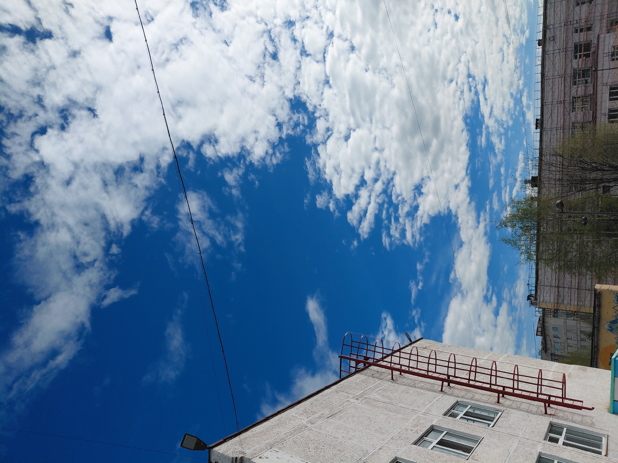 An outdoor, high-angle shot captures a bright blue sky filled with scattered white cumulus clouds, dominating the upper and left portions of the frame. The scene depicts an urban environment, likely in Nadym, Russia, under bright daylight. Several thin black wires or power lines traverse the sky diagonally and horizontally across the image.
In the foreground, a light-colored, multi-story building with a textured facade occupies the lower right quadrant. It features multiple rectangular windows with white frames. A prominent red metal ladder, equipped with safety hoops, is attached to the side of this building, extending diagonally upwards. A dark, rectangular street lamp is affixed to the building's top-left corner.
Further in the background, another multi-story building is visible on the right, largely covered in scaffolding and white protective netting, indicating ongoing construction or renovation. A small, dark figure, possibly a worker, can be seen on top of this scaffolding. A glimpse of a third building behind it reveals a colorful mural or graffiti. Sparse, light-green trees are visible between the buildings, suggesting spring or early summer. The weather is clear and sunny. No discernible text is visible in the image.