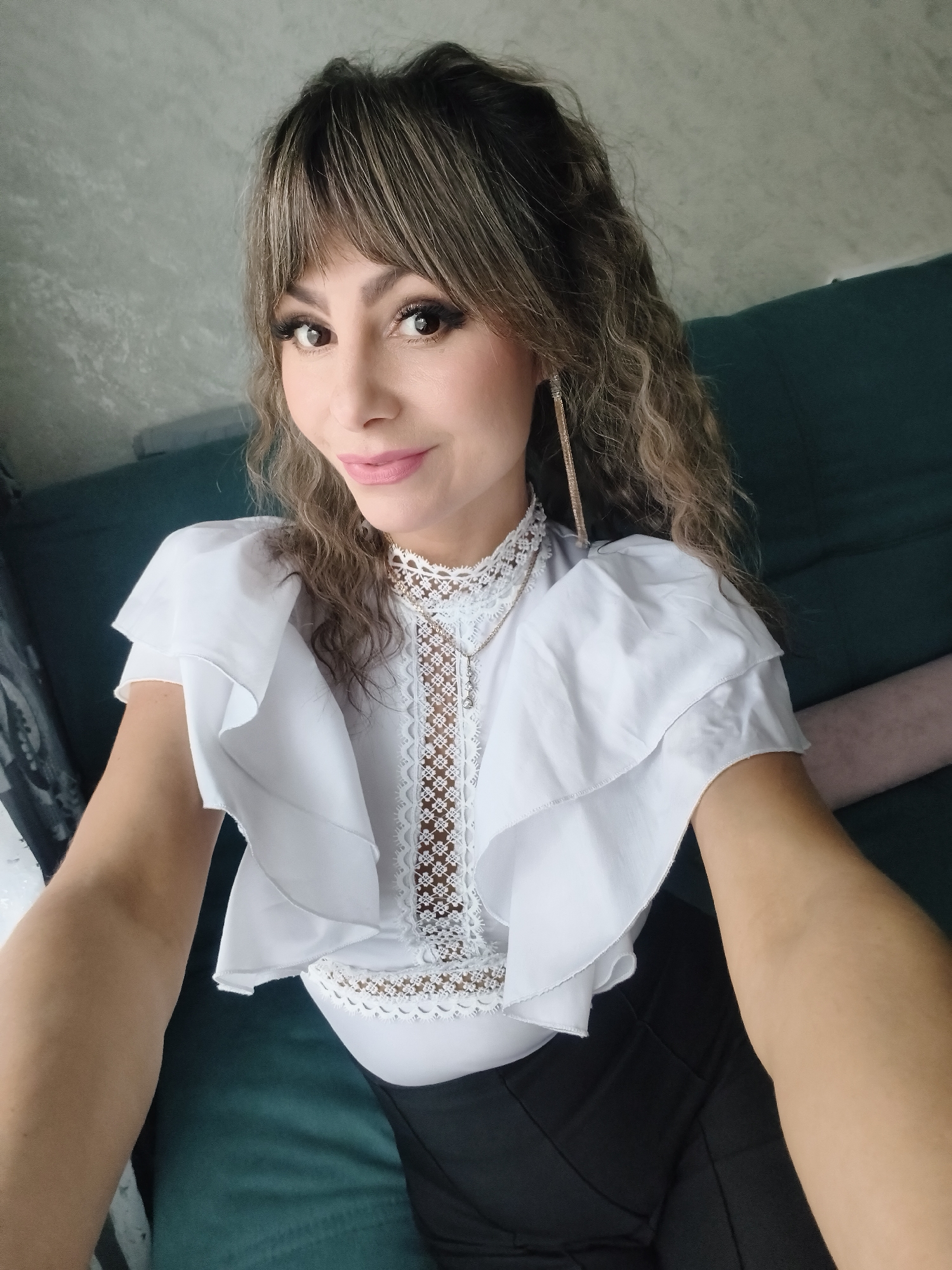 A medium-shot, eye-level selfie of a woman with curly brown hair and bangs is taken indoors, likely in Chaikovsky, Russia. She is wearing a white blouse with lace detailing and layered ruffled sleeves, paired with black pants. A long gold earring is visible. She is sitting on a dark green couch with a light pink cushion. The wall behind her is a light grey. The woman is looking at the camera with a slight smile.