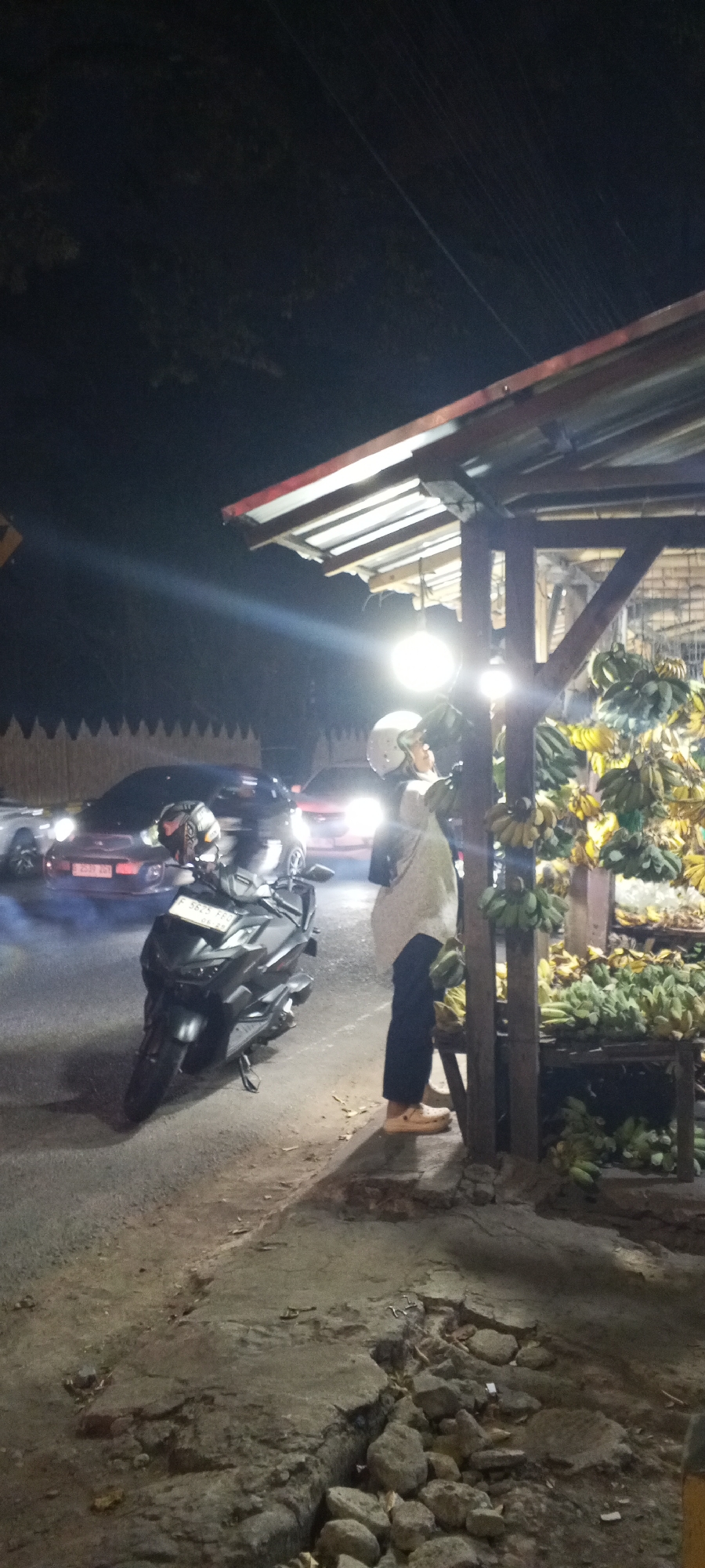 A nighttime scene on a street in Cibinong, Indonesia, shows a woman standing by a stall selling bananas. She is wearing a white hijab and a light-colored, long-sleeved top with dark pants and light-colored shoes. She appears to be looking at or interacting with the bananas. A large, dark-colored scooter is parked in front of her. The scooter has a license plate that reads "F 5825 FKG". Behind the woman and to her right, several bunches of bananas are displayed on wooden shelves under a simple roof structure supported by wooden posts. A bright light bulb hangs from the roof, illuminating the area. In the background, several cars are visible on the road, with their headlights on. One car, a red sedan, has its brake lights illuminated. The road appears to be asphalt and is uneven in the foreground, with cracked pavement and scattered rocks. A wooden fence with pointed tops is visible behind the cars. The sky is dark, indicating it is nighttime. The overall impression is of a roadside market operating late at night.