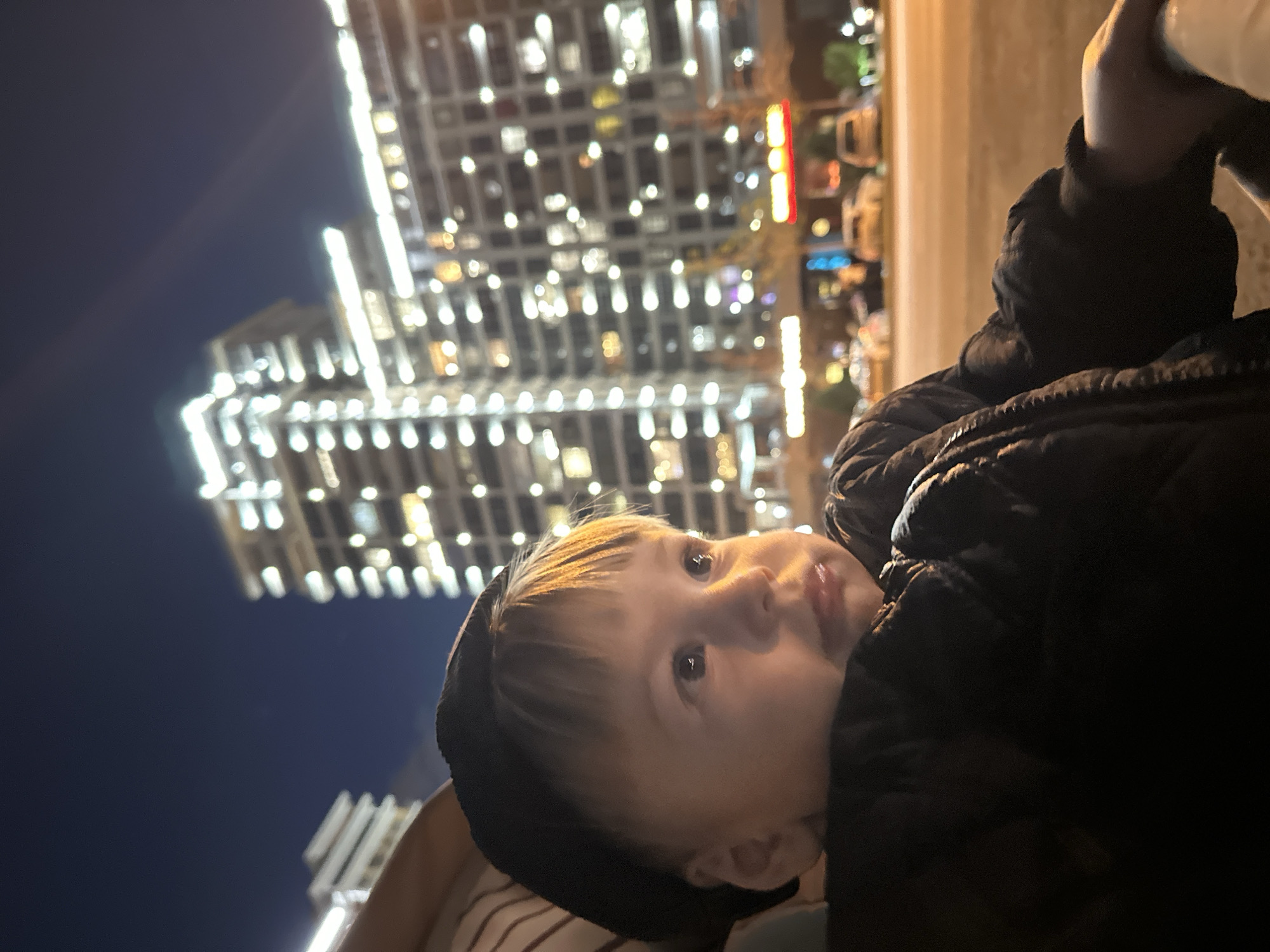 A close-up, slightly tilted, eye-level shot captures a young child wearing a dark-colored hooded jacket, with the hood pulled up. The child is looking upwards and slightly to the left of the frame. The background features tall buildings illuminated with numerous lights, typical of a cityscape at night. The buildings suggest a modern urban setting. This image appears to be taken in Kazan, Russia based on the building styles and lighting.