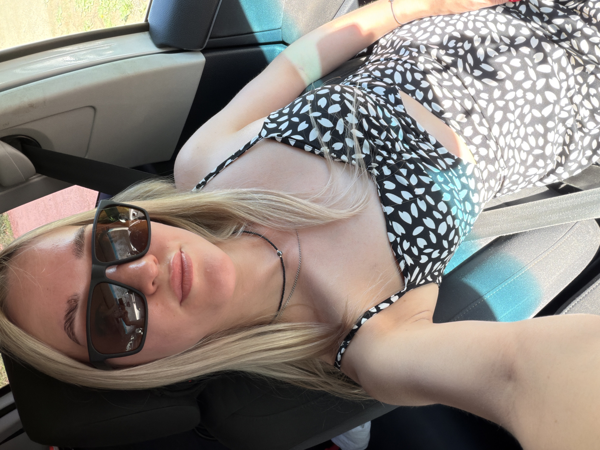 A woman lies in the passenger seat of a vehicle. She wears a black and white patterned dress and sunglasses. A seatbelt is visible across her body. The interior of the vehicle is light-colored with black accents. The location is unknown.