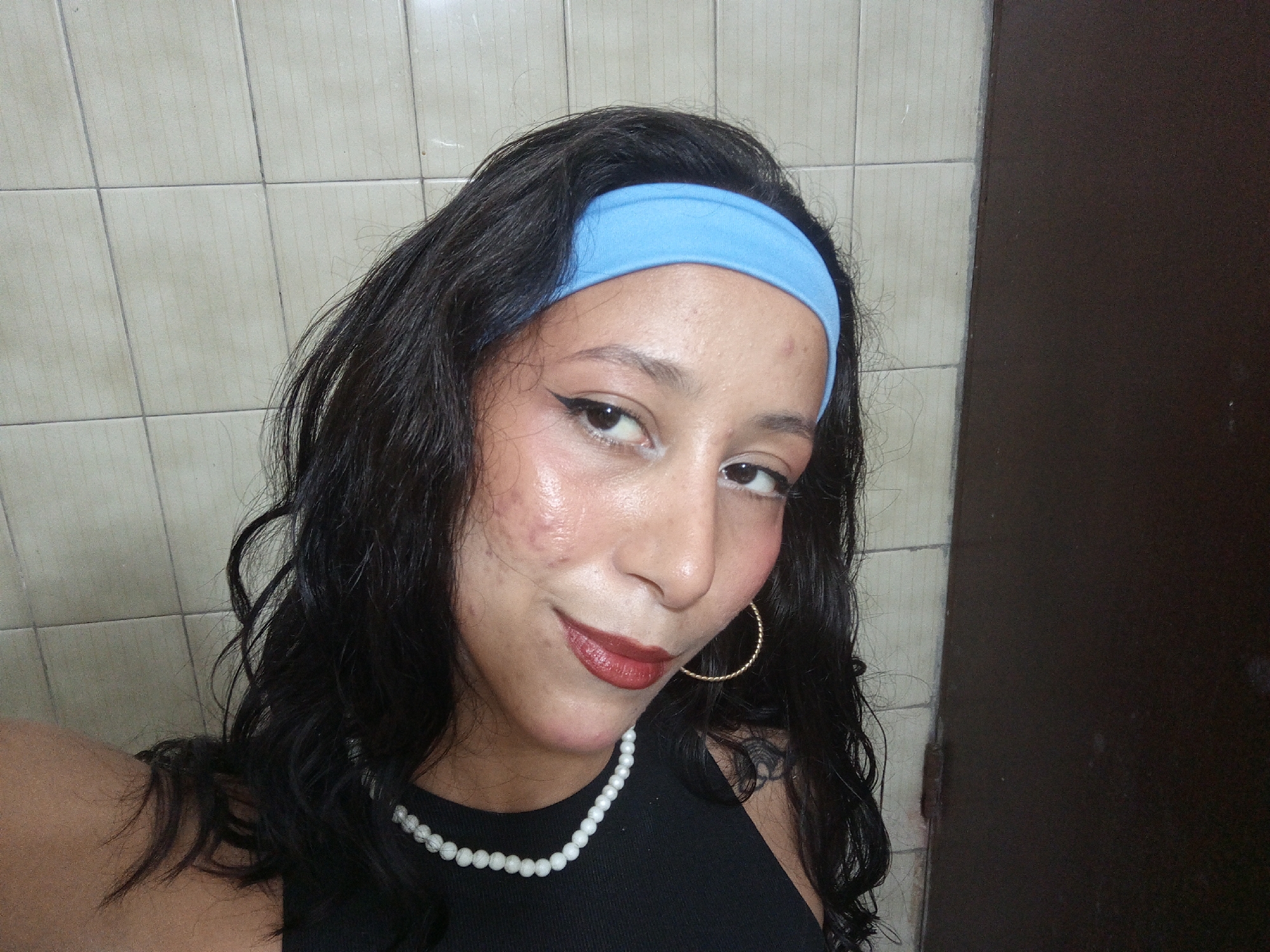 A young woman with dark, wavy hair and a light blue headband is centered in the frame. She is wearing eyeliner, lipstick, a gold hoop earring, and a white pearl necklace. She is wearing a black top. The background features a tiled wall and a dark wooden door. The location in the city of Porlamar, Venezuela cannot be determined based on the visual information provided.