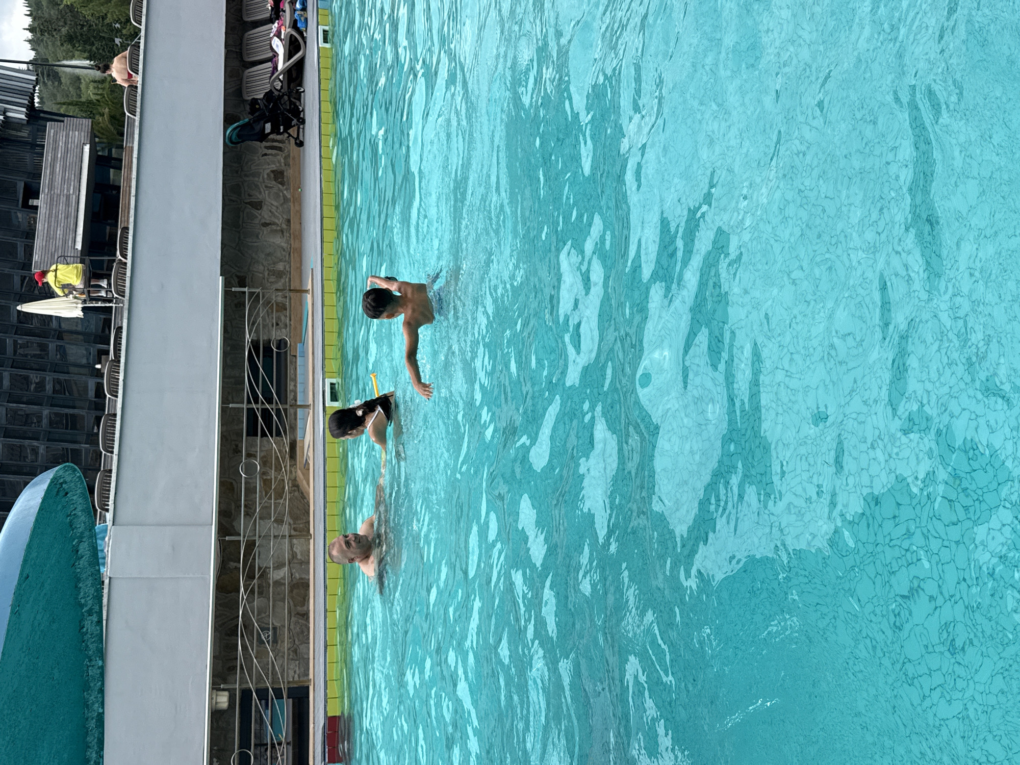 In a bright, turquoise swimming pool, three individuals are visible. A man with a shaved head is in the water, facing a young girl with dark hair tied back in a ponytail. The girl is wearing a black and white patterned swimsuit. Further to the right, a young boy with dark hair is swimming away from the camera, his right arm raised. The pool has a textured bottom visible through the clear water. In the background, above the pool, a grey wall with horizontal metal railings is present, behind which lies a stone-faced structure. Further in the distance, a modern building with a dark, angled roof is visible, with trees and a cloudy sky beyond. On the upper level of the building, a person wearing a yellow shirt and red hat sits on a chair, appearing to be a lifeguard. Several lounge chairs and a baby stroller are also visible on the deck of the pool. The scene appears to be at a public pool, possibly within the Массандровский поселковый совет, Ukraine.