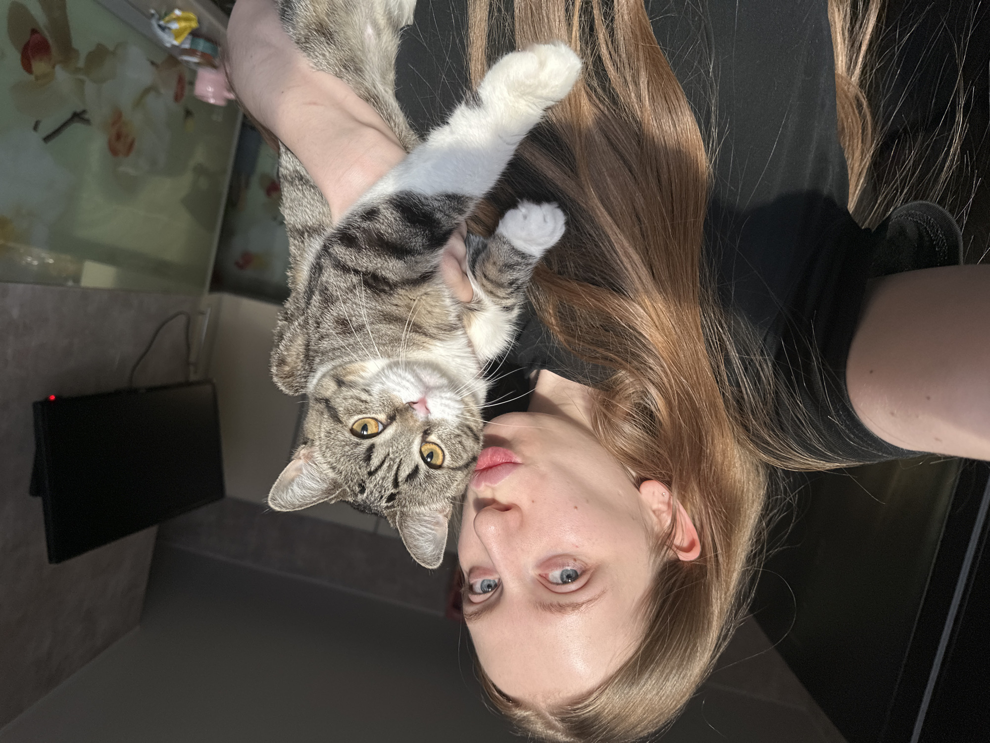 A female individual, appearing in her 20s with long, light brown hair and blue eyes, is depicted in a close-up selfie within an indoor residential setting in Chaikovsky, Russia. She wears a black short-sleeved top and a small stud earring in her left ear. She holds a tabby cat with distinct grey, brown, and black stripes, white chest and paws, and amber eyes. The female's lips are pursed in a 'kissing' gesture towards the camera, while the cat looks directly forward. Strong natural light enters from the left, illuminating the subjects. The background on the right features a dark grey, textured wall with a mounted black flat-screen television displaying a red power indicator light and visible cables. Below the television, on a surface, a light pink mug, a small container, and a crumpled yellow and white wrapper are present. Two framed pictures, likely depicting orchids with white and red/orange petals, are partially visible in the lower right background. The ceiling is dark.