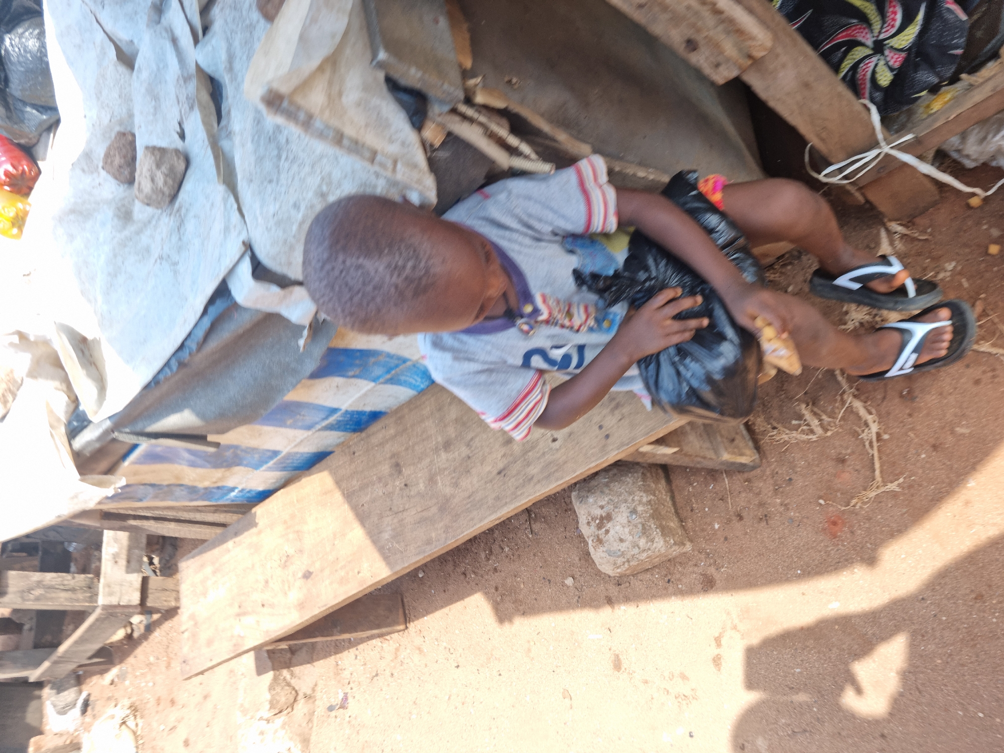 An overhead photograph captures a young child, likely a boy, resting or sitting on a makeshift surface composed of wooden planks and layered tarpaulins. The child is dressed in a light grey t-shirt with red and white striped short sleeves and dark-colored flip-flops with white stripes. He holds a black plastic bag with his left hand and a light-colored piece of food, possibly a snack or bread, in his right hand, appearing to be focused on it.
The immediate environment is cluttered with various materials. Below the child are several wooden boards and a blue and white striped tarpaulin. To the upper left, white or light-colored fabric or tarpaulin is scattered with a few small rocks. The ground is a reddish-brown dirt or sand. Strong, distinct shadows are cast across the scene, indicating bright daylight, likely midday or early afternoon. The setting appears to be an outdoor, informal, and possibly improvised area, characterized by the use of found materials and general debris. There is no legible text visible in the image. The child's facial expression is not clearly discernible due to the angle.