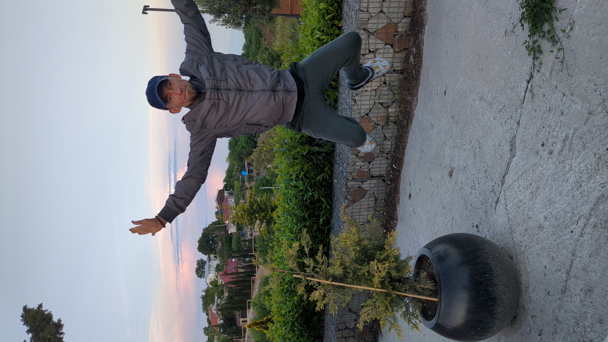 A man is suspended mid-air in front of a landscape. He wears a dark blue baseball cap, gray jacket, dark green pants, and patterned slip-on shoes. His arms are extended outward. Behind the man are trees, houses, and a gradient sky of pink, orange, and blue. A gabion wall is visible behind him, and a tree planted in a black pot. The location is Kiparisnoe, Ukraine.