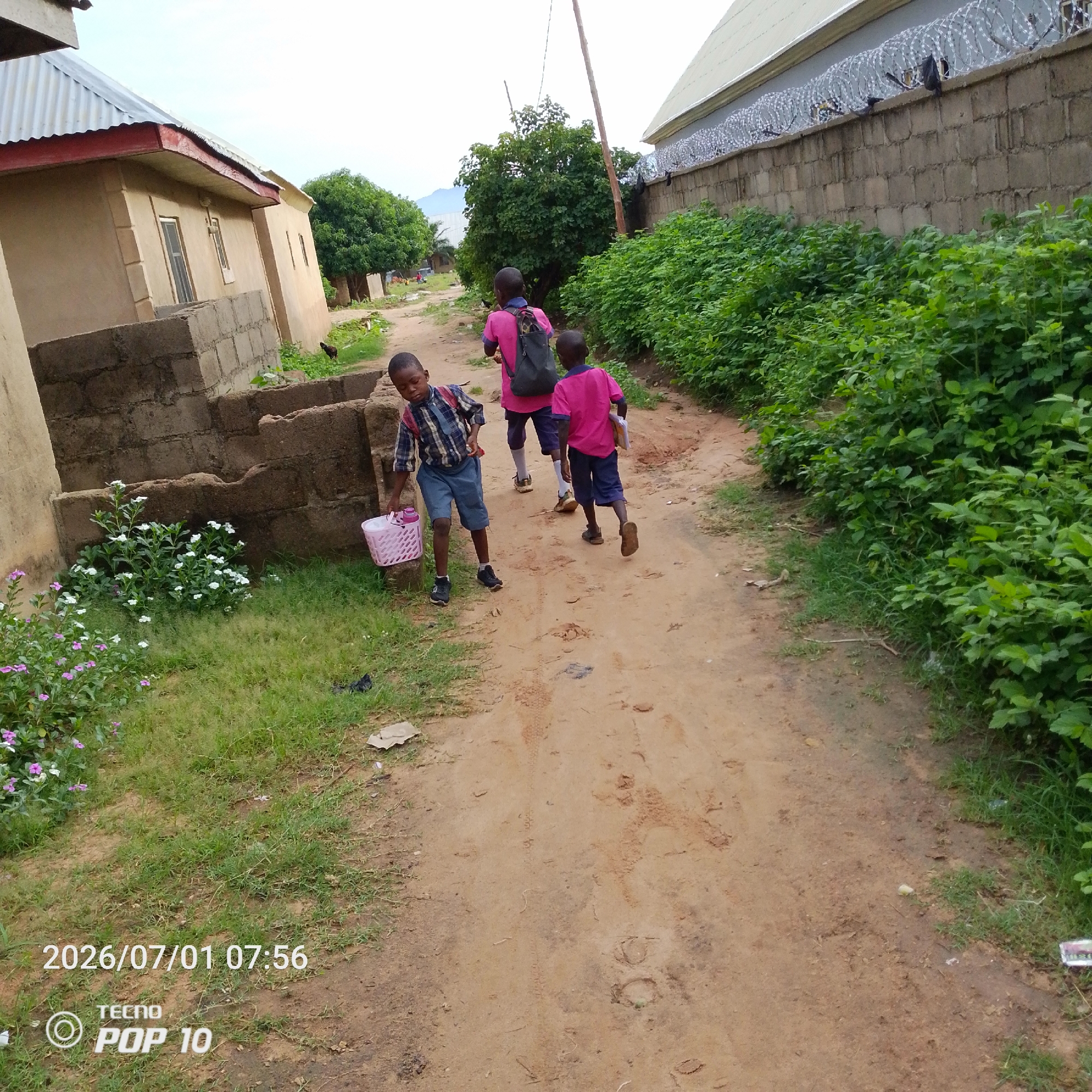 Three young boys are walking on a dirt path in Jalingo, Nigeria. The boy in the foreground, wearing a plaid shirt and blue shorts, carries a pink woven basket. Behind him, two other boys, both wearing pink shirts and dark shorts, walk ahead. The boy in the middle has a dark backpack. The path is flanked by vegetation on the right and buildings and more vegetation on the left. A brick wall topped with barbed wire runs along the right side of the path in the background. The sky is overcast. The timestamp "2026/07/01 07:56" and "TECNO POP 10" are visible in the lower left corner.