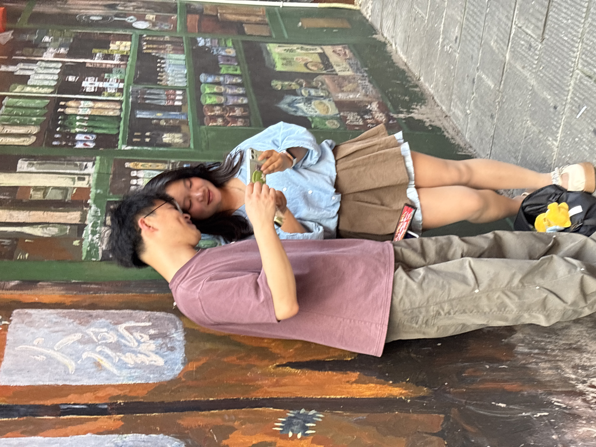 In Yuzhong District, China, a young East Asian couple stands next to a mural-covered wall. The man, wearing a maroon t-shirt and olive-green pants, looks down at his phone, which he holds in his right hand. The woman, dressed in a light blue button-down shirt and a brown pleated skirt over a white hem, gazes at the phone in her hands. The mural on the wall depicts various items on shelves, resembling a shop. To the left of the couple, another section of the mural shows what appears to be Chinese calligraphy. The ground is paved with gray stone tiles. A black backpack with a yellow charm is visible at the woman's feet. The lighting suggests it is daytime.