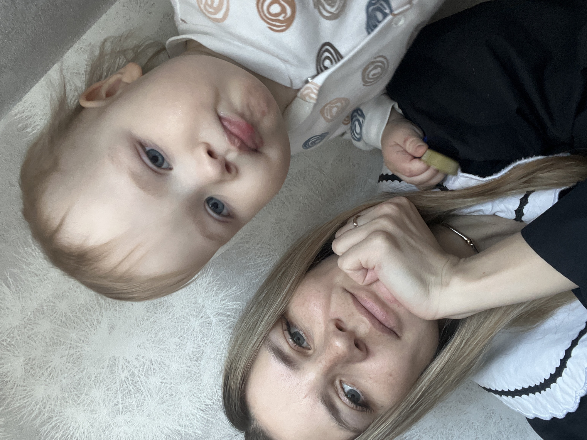 A close-up, diagonal selfie captures an adult female and a baby indoors in Chaikovsky, Russia. The woman, positioned in the lower-left, has fair skin, light blue-grey eyes, long straight blonde hair, and visible freckles on her nose and cheeks. She wears a black garment with a prominent white scalloped collar and a thin metallic choker. A ring is visible on her right ring finger. Her right hand supports her chin, and she looks directly at the camera with a slight smile. The baby, positioned in the upper-right, has fair skin, blue eyes, and light hair, also looking directly at the camera with a neutral, slightly pursed expression. The baby wears a light-colored long-sleeved outfit or sleepsuit patterned with brown, blue, and grey abstract circular motifs; the front appears partially unzipped. The baby's left hand holds a small, yellowish-green object, resting on the woman's shoulder. The background is a light-toned wall with a subtle, repeating starburst or dendritic patterned wallpaper.
