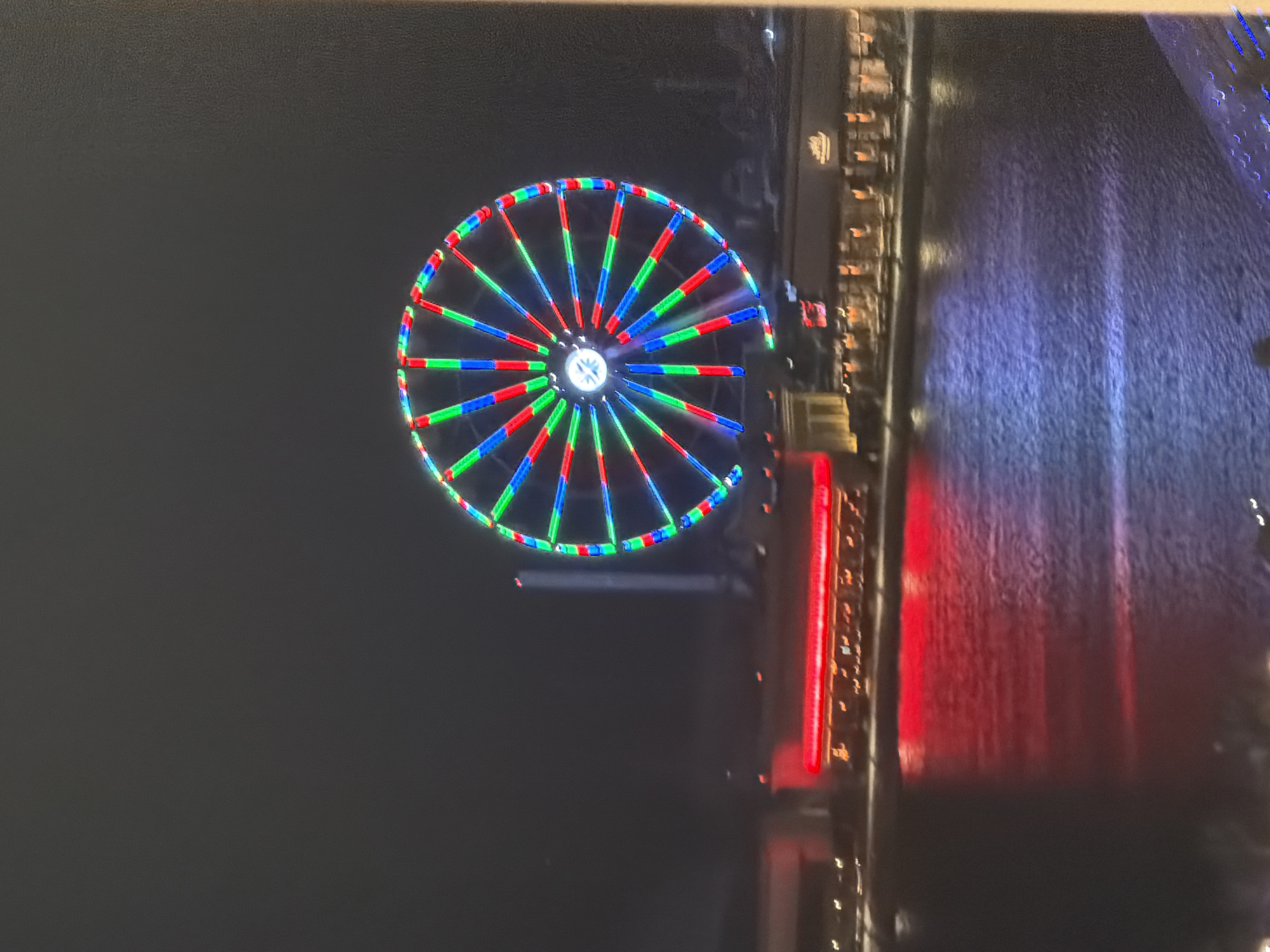 The image depicts a nighttime scene in the city of Unknown, Unknown, featuring a prominent Ferris wheel and illuminated waterfront buildings. A large Ferris wheel, located slightly right of center in the mid-ground, is brightly lit with alternating red, green, and blue LED segments on its spokes and outer rim; its central hub emits a white or light blue glow. Below the Ferris wheel, a body of water occupies the lower half of the frame, displaying rippled reflections of the surrounding lights.
On the left side of the image, a building features a distinct horizontal band of red illumination along its upper facade, which is strongly reflected in the adjacent water. To the right, another commercial building or complex is visible, illuminated with warmer, yellow-orange lights emanating from windows or interior spaces. A partial sign on this rightmost building appears to read "MELT" or similar. The dark night sky fills the upper portion of the frame, devoid of visible stars or clouds. The absence of discernible individuals indicates no specific human activity.