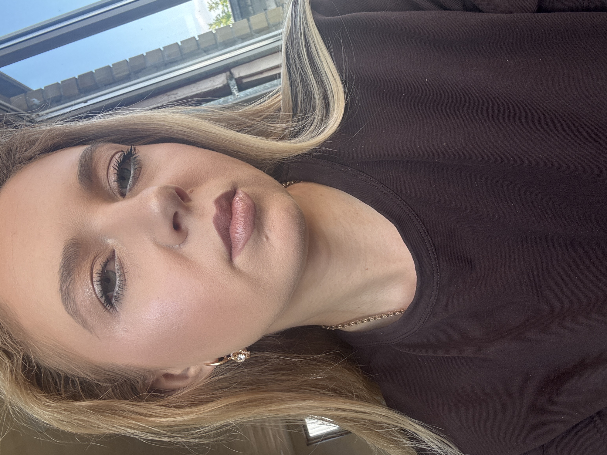 A young woman with blonde hair, wearing makeup and a brown shirt, is pictured from the chest up. She is wearing a gold hoop earring with a clear stone, and a gold chain necklace. She is looking at the camera. The background consists of a window with a blue sky and some vegetation visible outside.