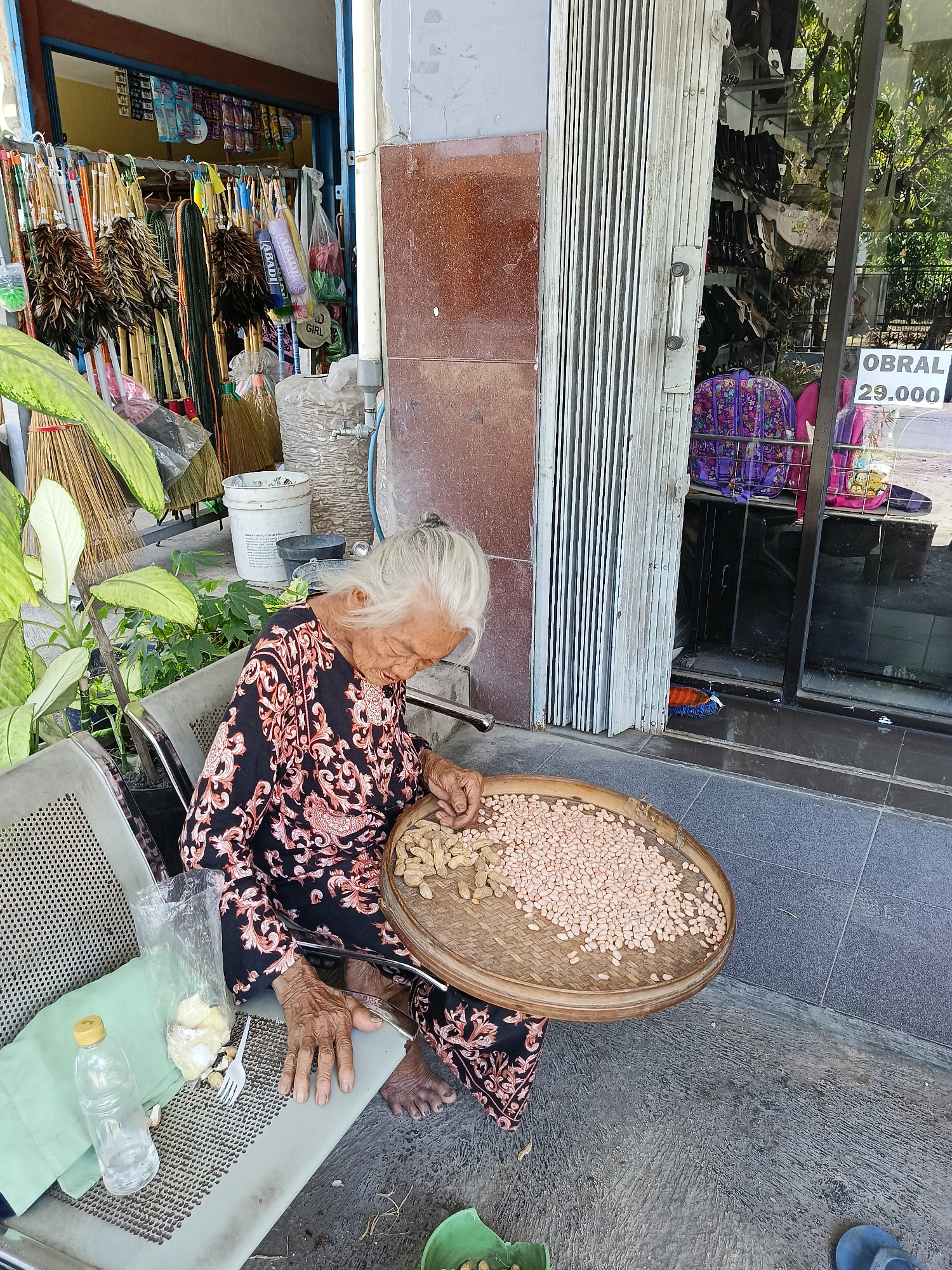 An elderly woman with white hair sits on a metal chair in front of a storefront in Manisrenggo, Indonesia. She is wearing a black and orange patterned batik dress and is looking down at a large, circular woven tray filled with peanuts, which she appears to be sorting or processing with her hands. Next to her on the chair is a plastic bag containing some food items, a white plastic fork, and a clear plastic water bottle. Behind her, various items are displayed outside the storefront, including brooms, feather dusters, and other goods. A metal roll-up door partially covers the entrance to the store. The ground is paved with gray tiles. In the window of the store, a sign reads "OBRAL 29.000".