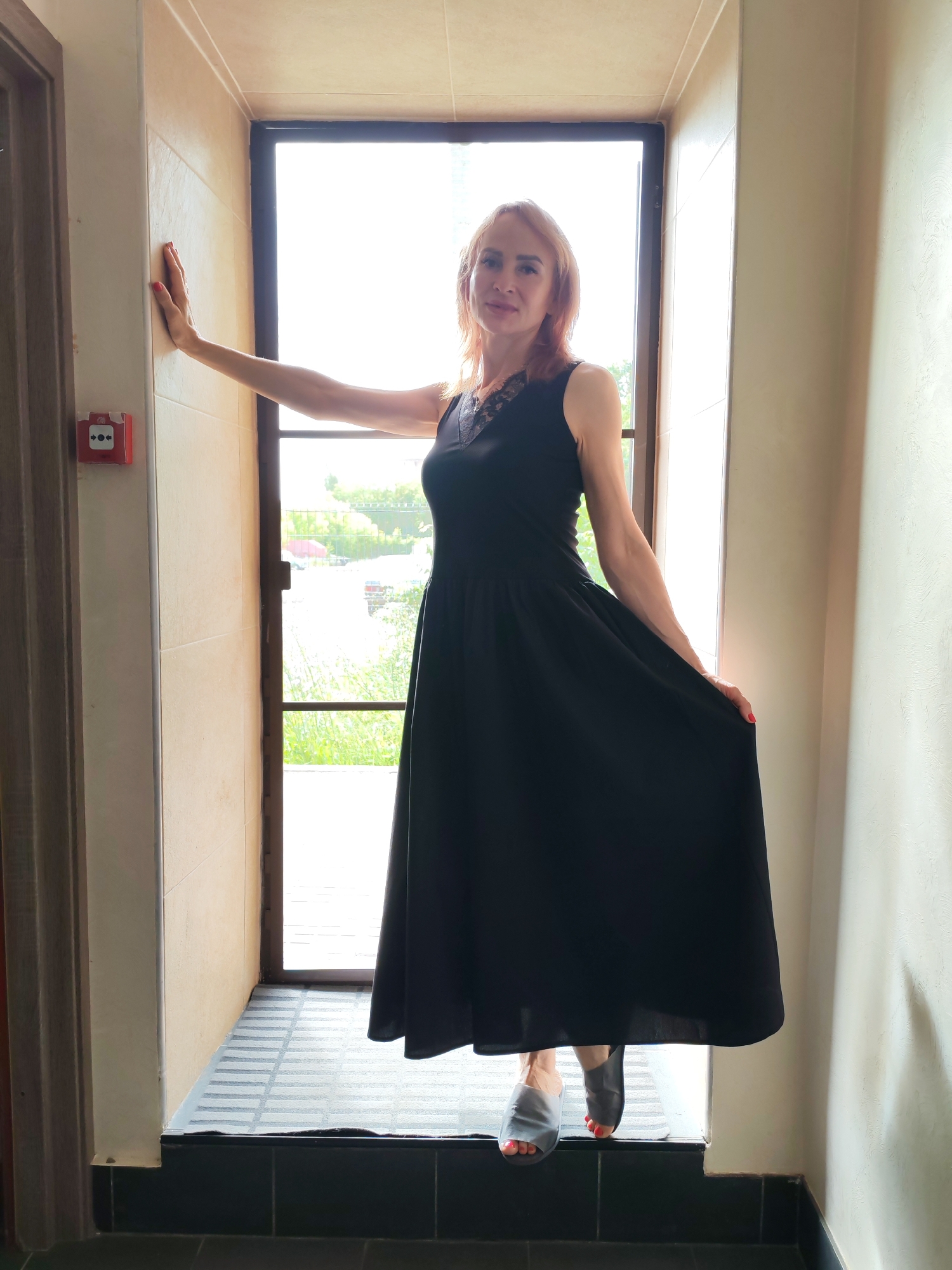 A woman with light skin and reddish-brown hair, wearing a black sleeveless dress with lace detailing at the neckline and gray slippers, stands in a doorway. She is leaning against a wall with her left hand and holding the skirt of her dress with her right hand. The background shows a large window with a view of greenery and a fence. The woman is positioned centrally within the frame, illuminated by natural light coming from the window. The surrounding environment appears to be indoors, with tiled walls and a tiled floor. A red fire alarm pull station is visible on the left wall. The location is identified as Yoshkar-Ola, Russia, though there are no specific visual cues within the image to confirm this geographical detail.
