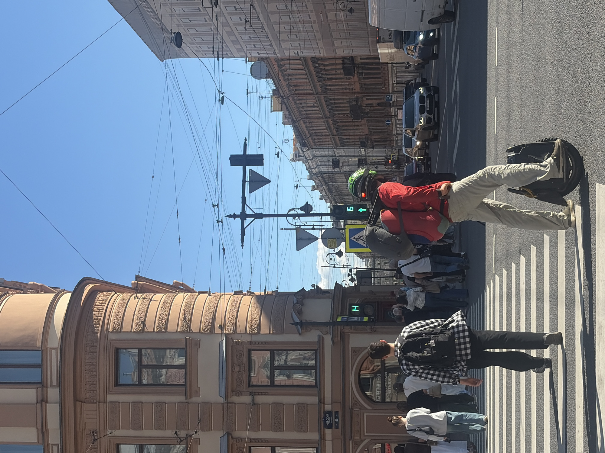 The image depicts a street scene in Saint Petersburg, Russia, likely on Nevsky Prospekt, as indicated by a street sign. The foreground features a pedestrian crossing where individuals are walking. A man is prominently positioned on the crossing, riding a self-balancing unicycle, wearing a red jacket, light-colored pants, a helmet, and a backpack. Behind him, other pedestrians are also crossing the street, including a man in a checkered shirt with a backpack. In the background, a row of multi-story buildings with classic architectural details lines the street. Traffic, including cars and a white van, is visible on the road. Overhead, a complex network of power and tram wires crisscrosses the sky, with traffic lights and street signs also present. The sky is clear and blue, suggesting a sunny day.