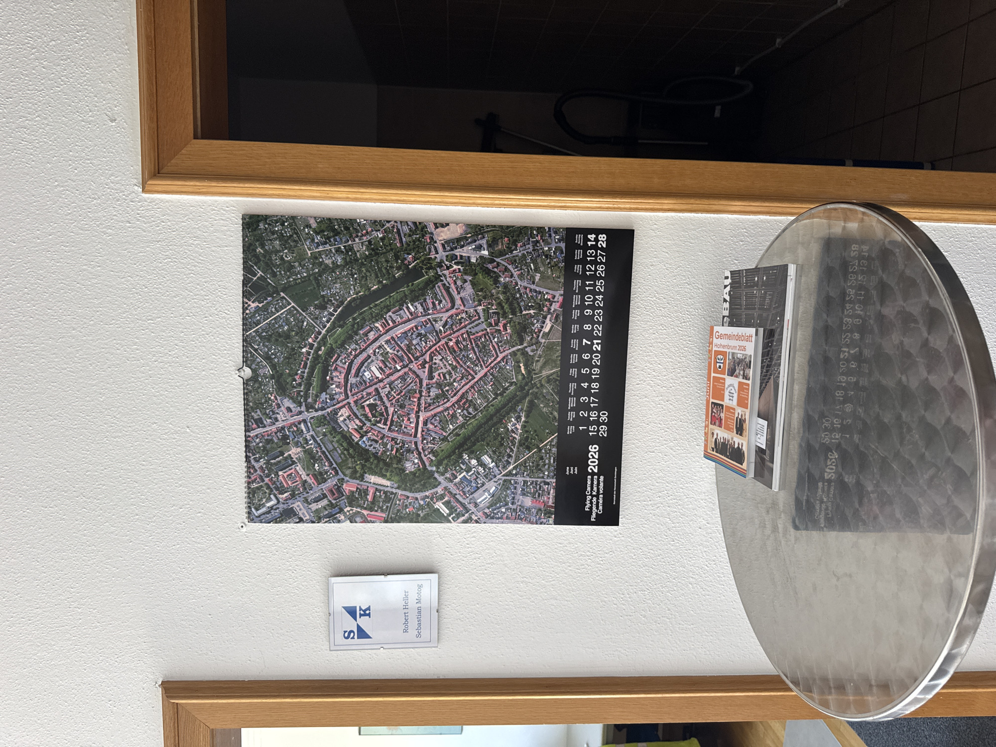 This image shows a close-up interior shot of a wall with a calendar hanging on it, and a round metal table in the foreground. The calendar features an aerial view of a circular town layout with buildings and greenery. The calendar is for June 2026 and is titled "Flying Camera" in English, "Fliegende Kamera" in German, and "Camera volante" in Italian. Below the calendar, the text "Gemeinde Hohenbrunn" indicates the location is Hohenbrunn, Germany. On the table, there are two magazines or pamphlets. The wall has a textured finish, and there is a wooden door frame on the right side of the image, opening into a dark room with tiled walls. A vacuum cleaner is visible in the dark room.
