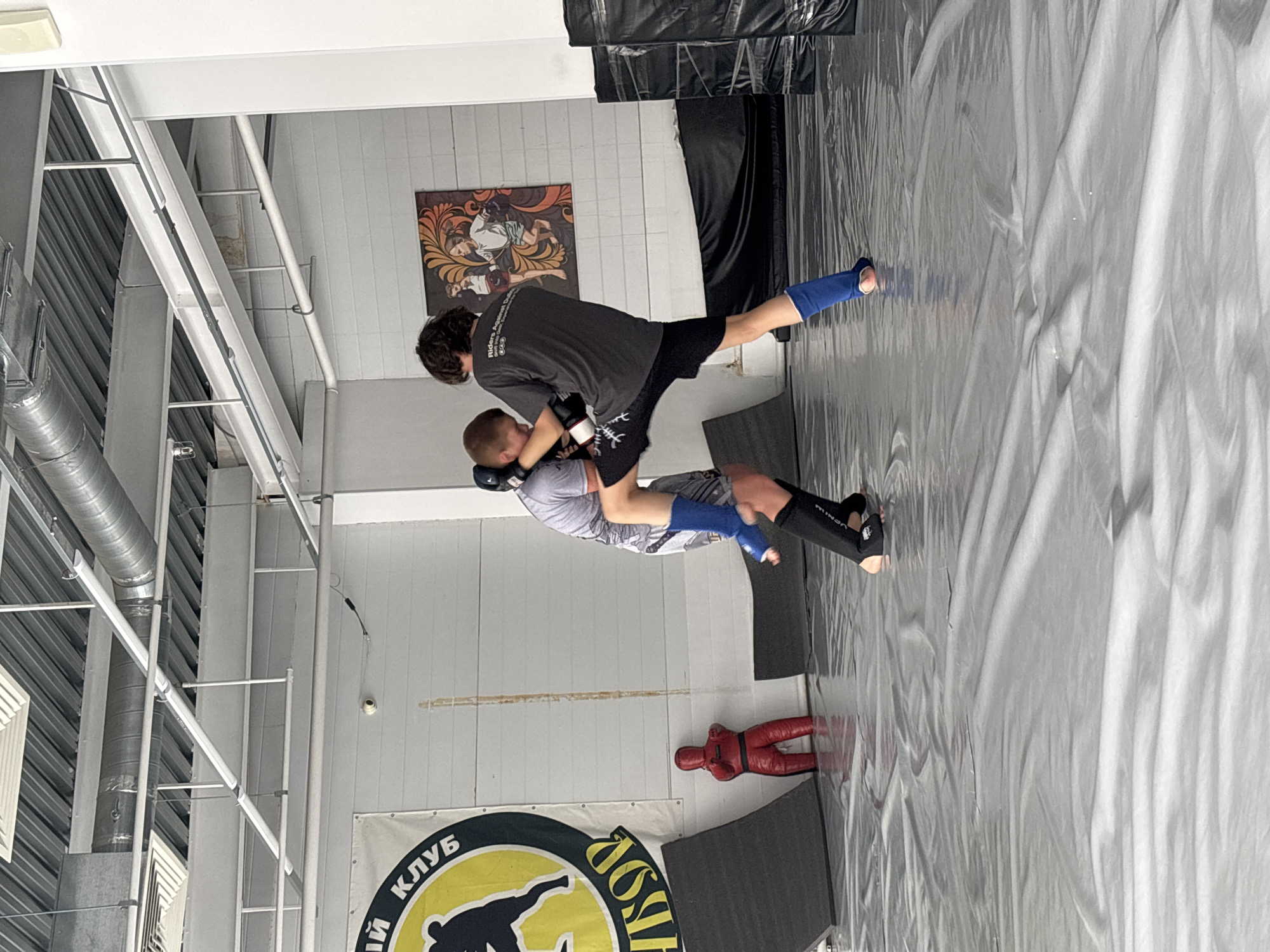 The image shows two individuals engaged in a grappling activity in a gym setting. One person, wearing a dark gray shirt and black shorts, has the other person in a hold. The second individual is wearing a light gray patterned shirt and gray shorts. Both are wearing protective gear on their hands and legs. They are on a gray mat in a room with a banner with a logo including the words "Пермский Клуб" (Perm Club). There is a poster on the wall. A grappling dummy is near the banner. The location is likely in the city of Perm, Russia based on the text on the banner.