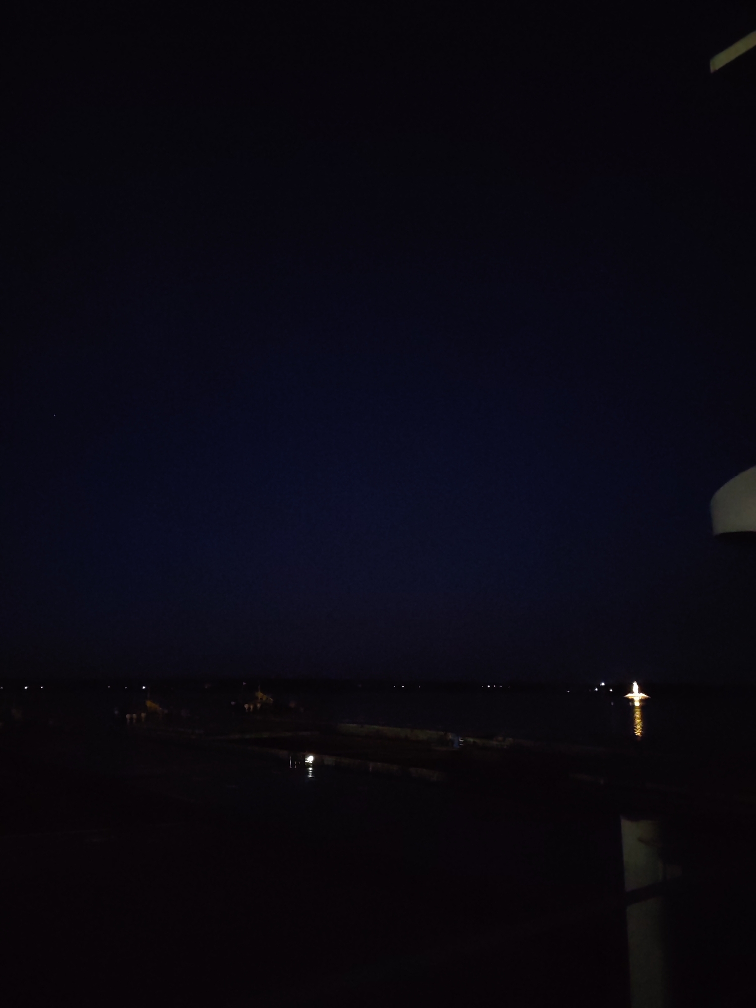The image shows a night view in Rybinsk, Russia. The sky is a deep blue, verging on black. In the distance, there is a body of water reflecting light from distant points. The water appears calm. The shoreline is dark, with small points of light scattered along it. To the right, there is part of a structure, potentially a column, that is out of focus and contributes to the darkness in the foreground. Overall, the scene is dimly lit, creating a sense of stillness and solitude.